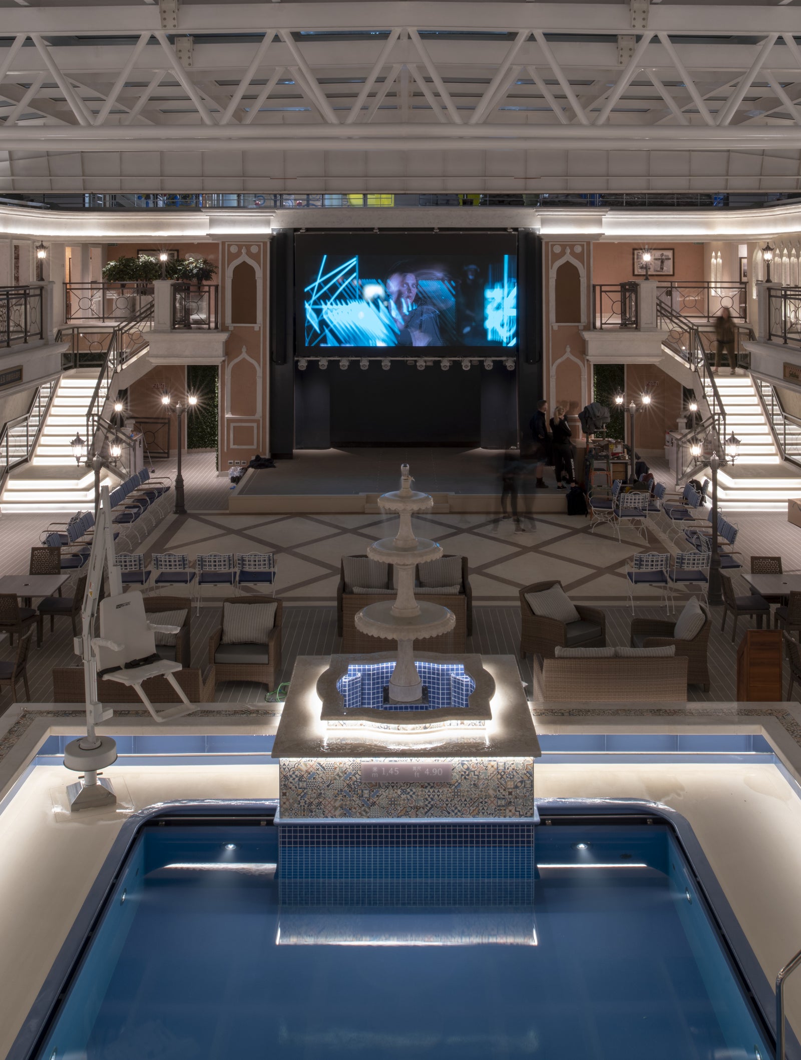 Fincantieri | Group - Business - Products - Cruise Ships - Costa Venezia: Spacious luxury cruise ship pool deck illuminated at night with elegant lights, central fountain, pool, deck chairs, refined architecture and large entertainment screen, setting designed for sophisticated relaxation and leisure.