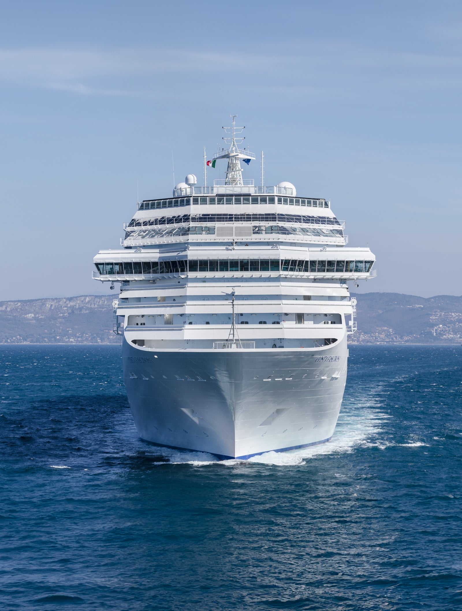  Fincantieri | Group - Business - Products - Cruise Ships - Costa Fascinosa: Massive white cruise ship in operation sailing forward on calm open waters, distant hills in background under clear blue sky, design includes multiple decks and visible flag at top.