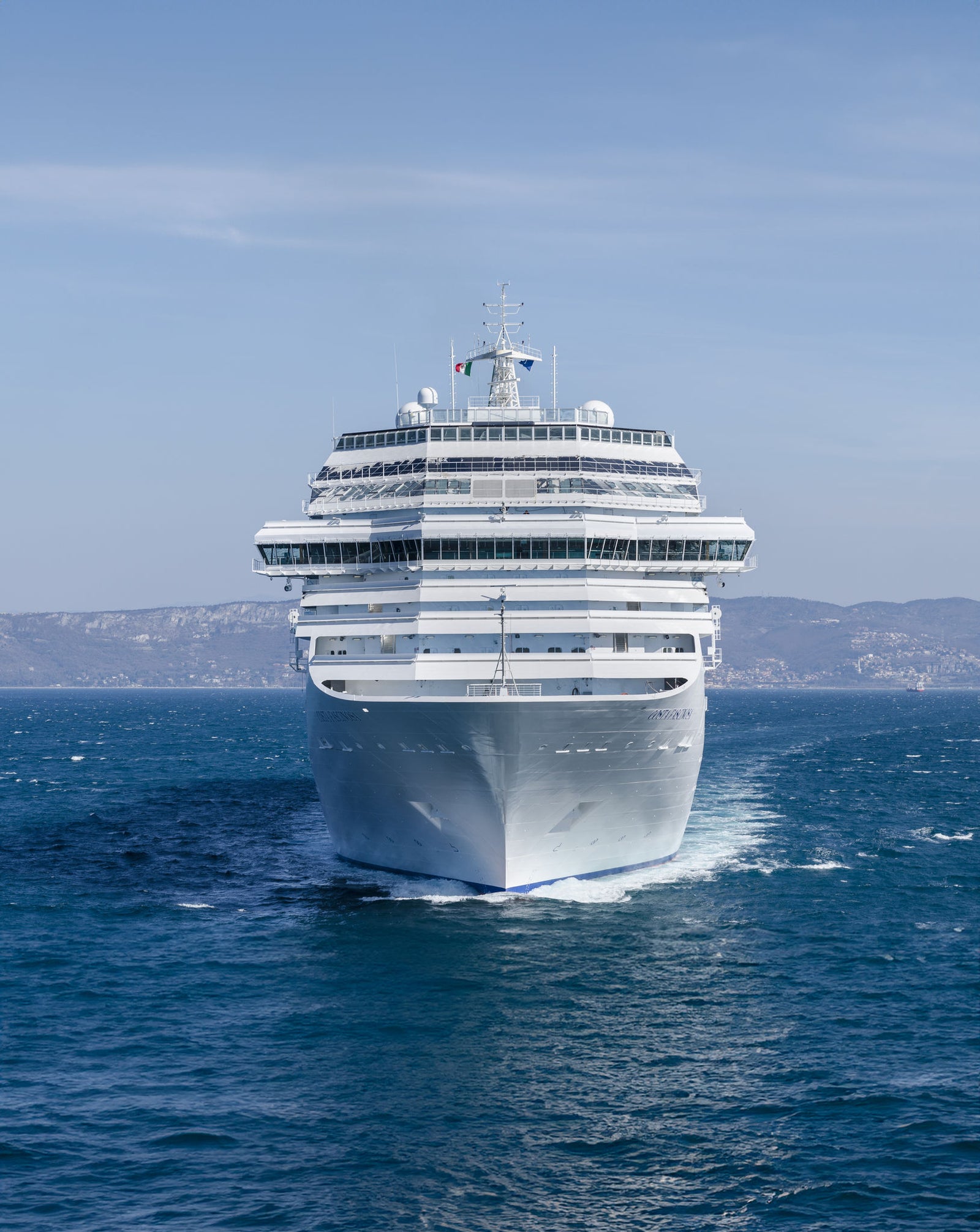 Fincantieri | Group - Business - Products - Cruise Ships - Costa Fascinosa: Massive white cruise ship in operation sailing forward on calm open waters, distant hills in background under clear blue sky, design includes multiple decks and visible flag at top.