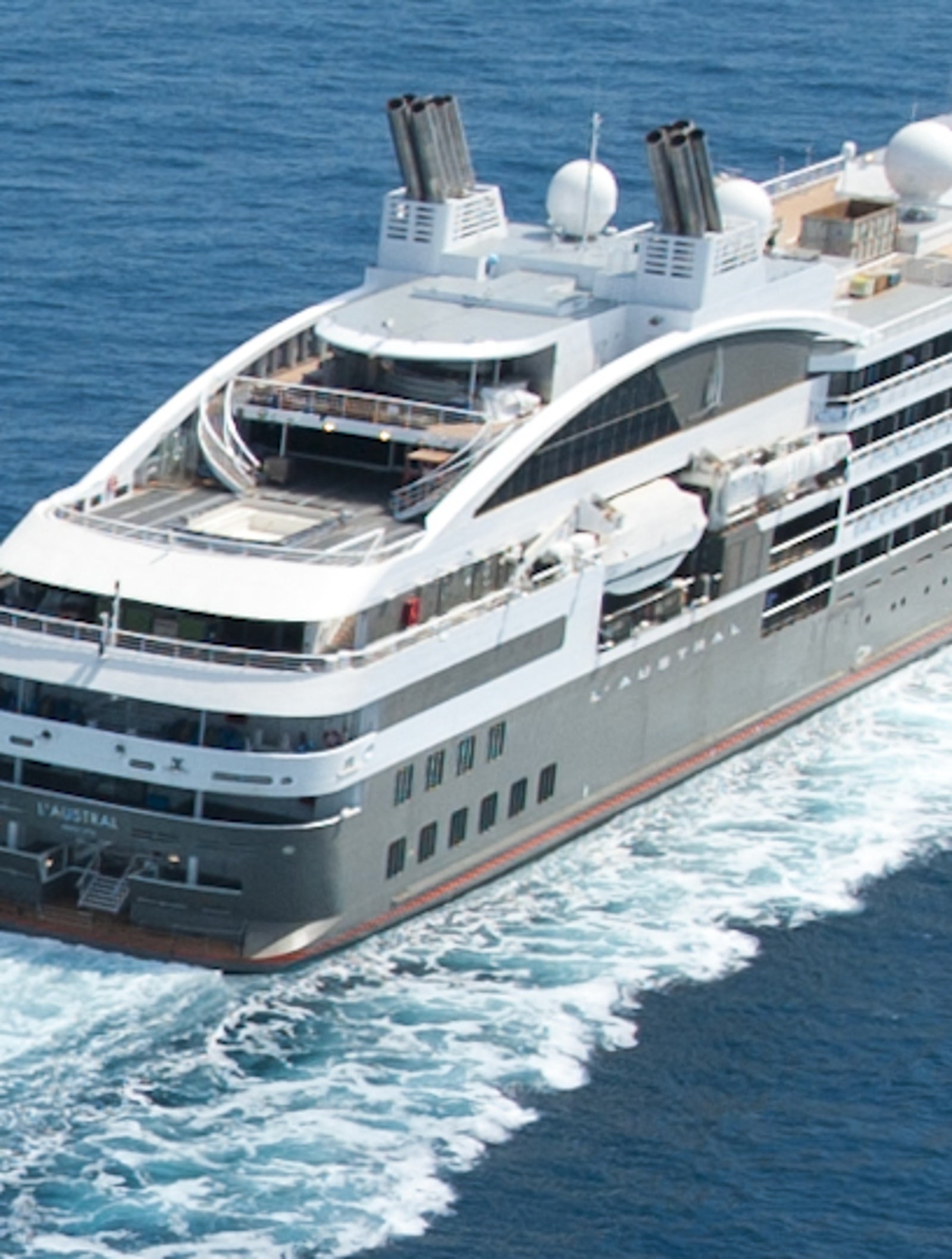  Fincantieri | Group - Business - Products - Cruise Ships - L'Austral: Large modern cruise ship in operation navigating calm deep blue open sea waters, visible wake trailing behind emphasizes movement and speed, clear sky and expansive ocean create serene and tranquil atmosphere.