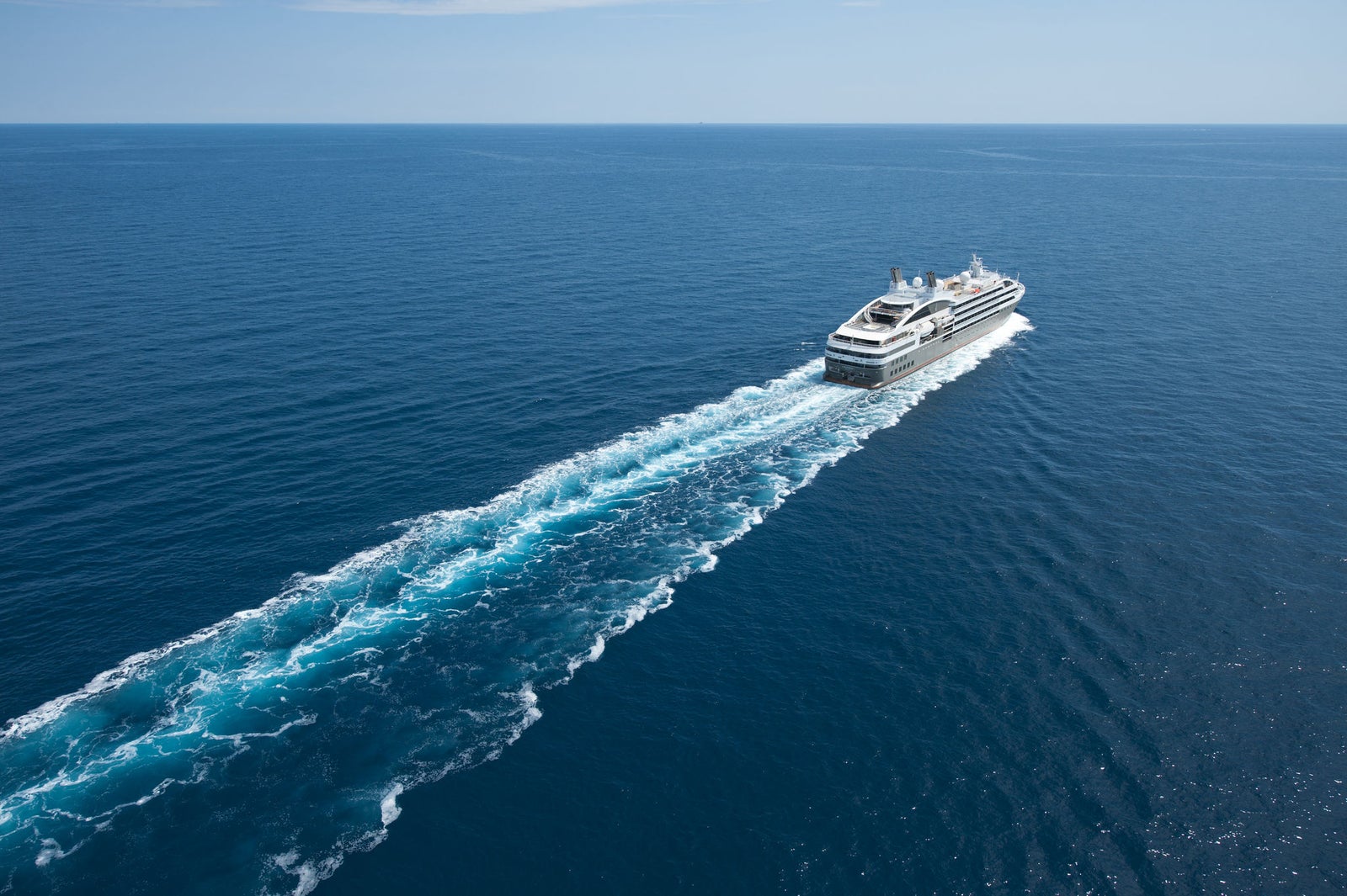 Fincantieri | Group - Business - Products - Cruise Ships - L'Austral: Large modern cruise ship in operation navigating calm deep blue open sea waters, visible wake trailing behind emphasizes movement and speed, clear sky and expansive ocean create serene and tranquil atmosphere
