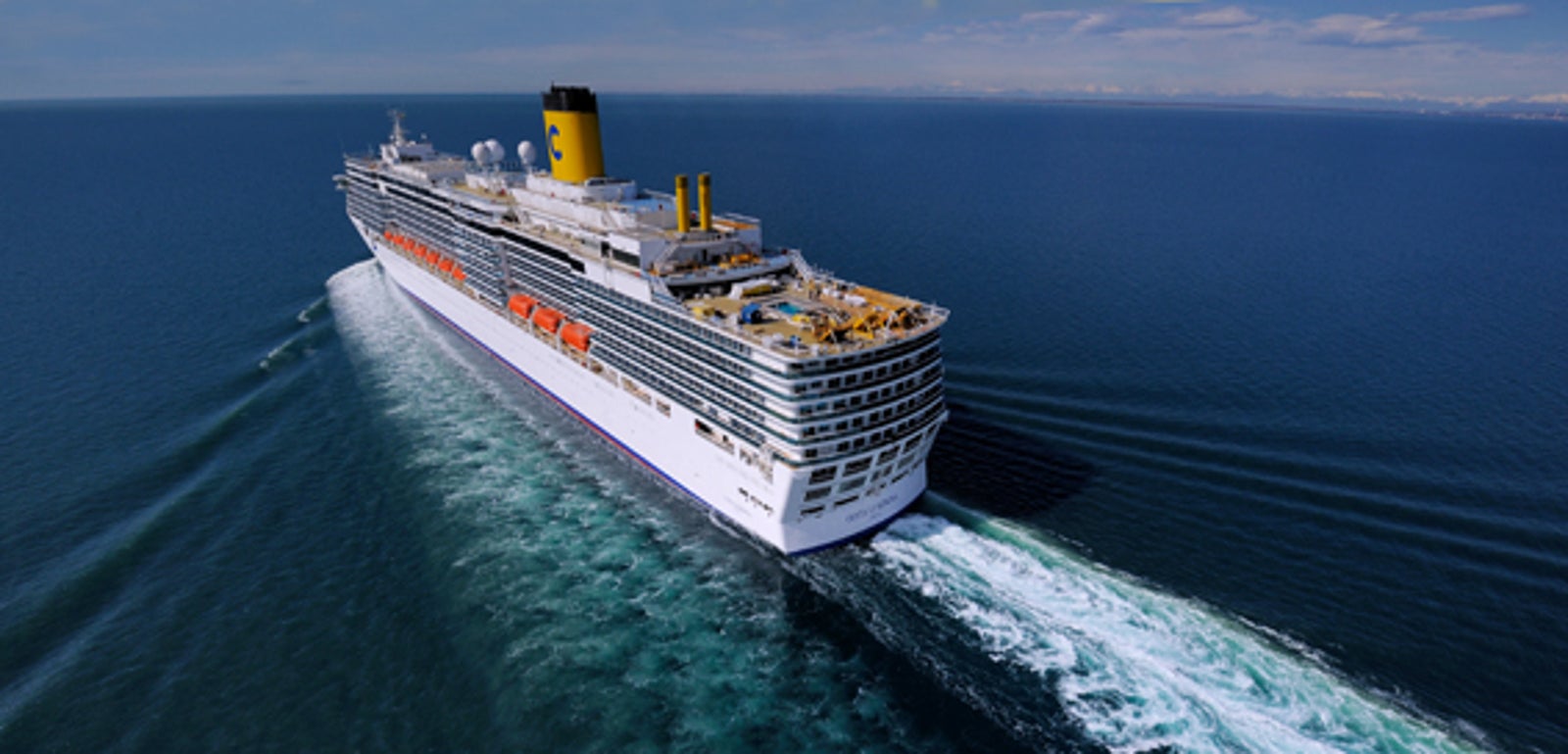 Fincantieri | Gruppo - Business - Prodotti - Navi da Crociera - Costa Luminosa: Grande nave da crociera bianca operativa con camino giallo naviga su oceano aperto, scia di schiuma bianca visibile, circondata da acque blu calme sotto cielo limpido, più ponti e scialuppe lungo i lati, indicano capacità per passeggeri.