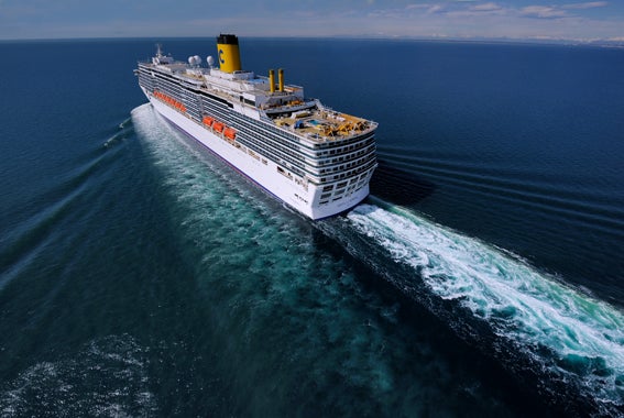  Fincantieri | Gruppo - Business - Prodotti - Navi da Crociera - Costa Luminosa: Grande nave da crociera bianca operativa con camino giallo naviga su oceano aperto, scia di schiuma bianca visibile, circondata da acque blu calme sotto cielo limpido, più ponti e scialuppe lungo i lati, indicano capacità per passeggeri.