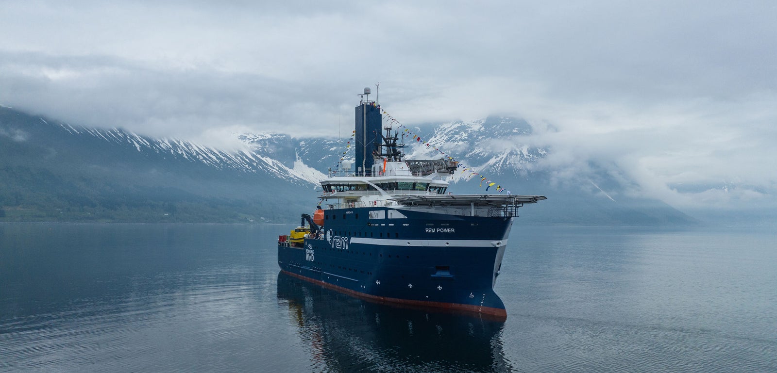 Prodotto - Offshore Support Vessel Rem Power: Vista laterale della nave operativa su acque calme, scafo blu con testo REM POWER visibile, sfondo montuoso con cime innevate e nuvole basse.