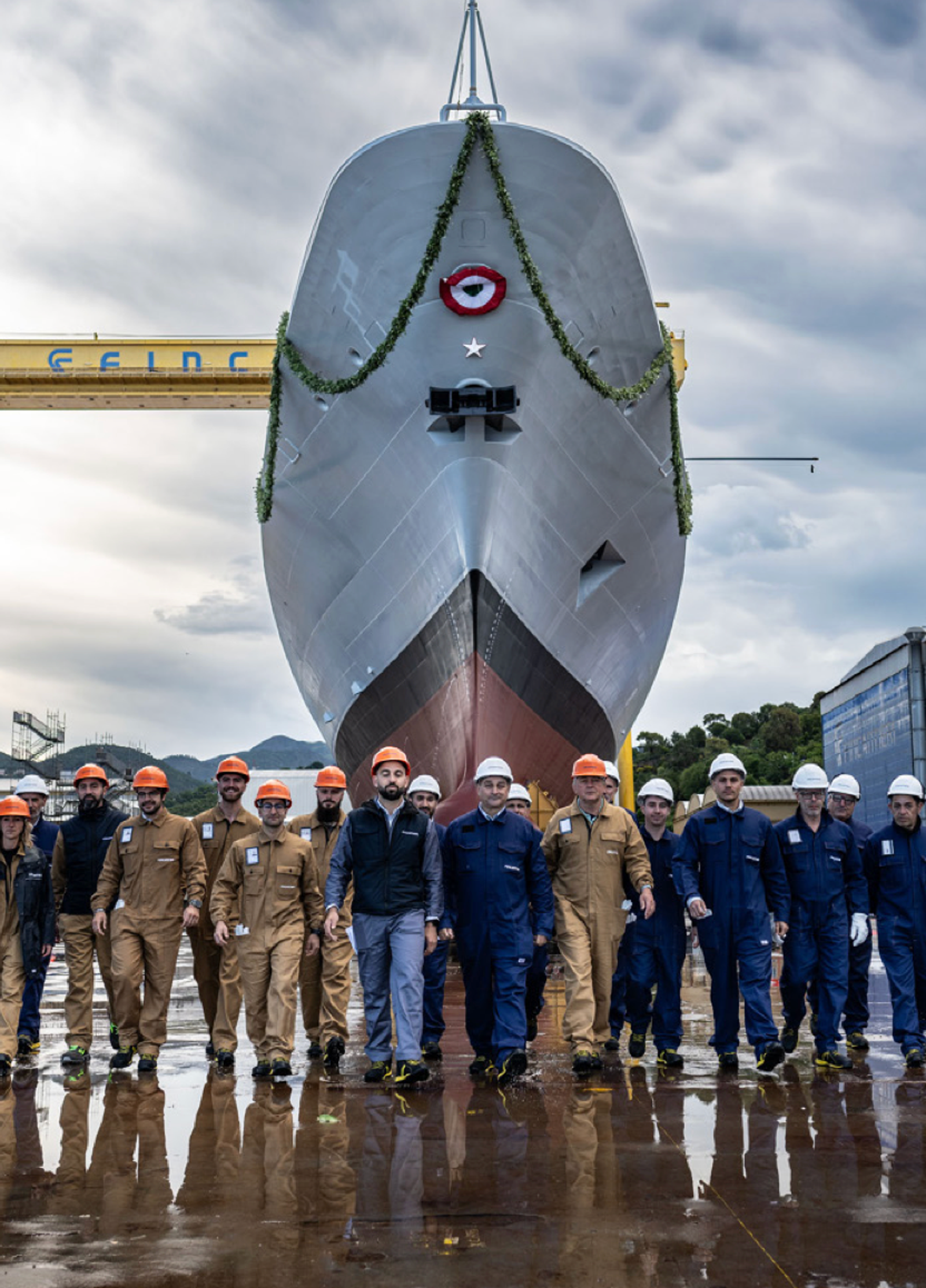Fincantieri | Persone - Inclusione Vivere Fincantieri: Gruppo di operai di cantiere navale in posa davanti a una nave durante la cerimonia di varo, nave decorata con ghirlanda verde sotto gru gialla CIMC, operai con caschi e divise di sicurezza, pavimento bagnato e cielo nuvoloso.