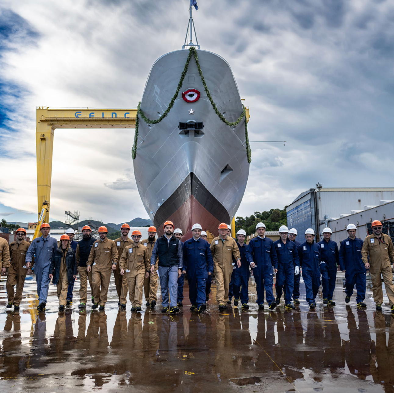 Fincantieri | Investing in Fincantieri: A group of shipyard workers poses in front of a large vessel during the launching ceremony. The setting is an industrial shipyard with cranes and buildings visible in the background. The workers wear uniforms and safety equipment, including helmets. The ship is decorated for the occasion, and the scene is captured under a partly cloudy sky.