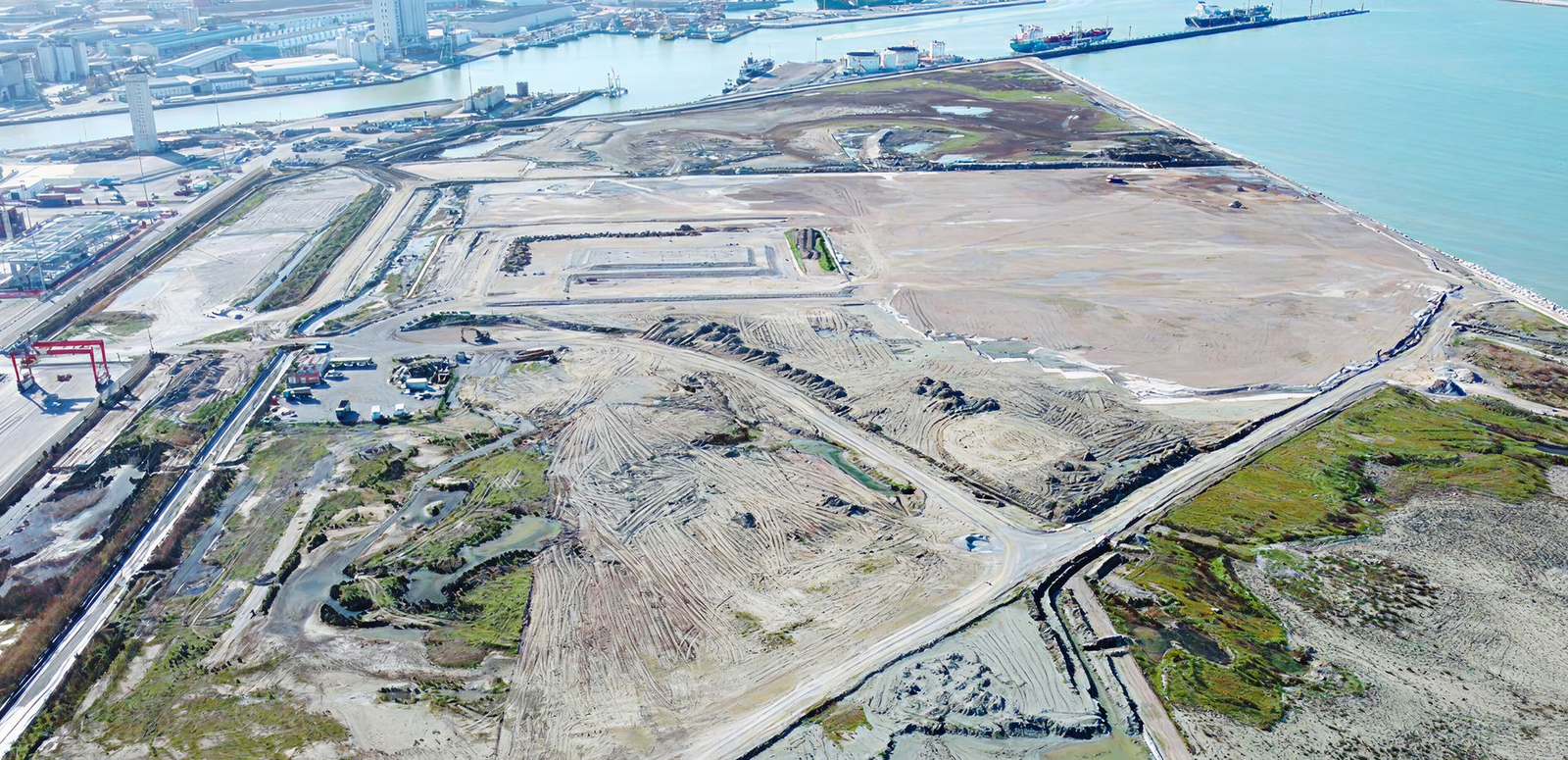 Fincantieri | Group - Business - Products - Maritime/Hydraulic Works - Coastal construction aerial view: Expansive aerial view of construction site near coastline; cleared land, industrial equipment, and surrounding water bodies visible; docks and ships in background indicate proximity to port, scene highlights development activity in coastal area.