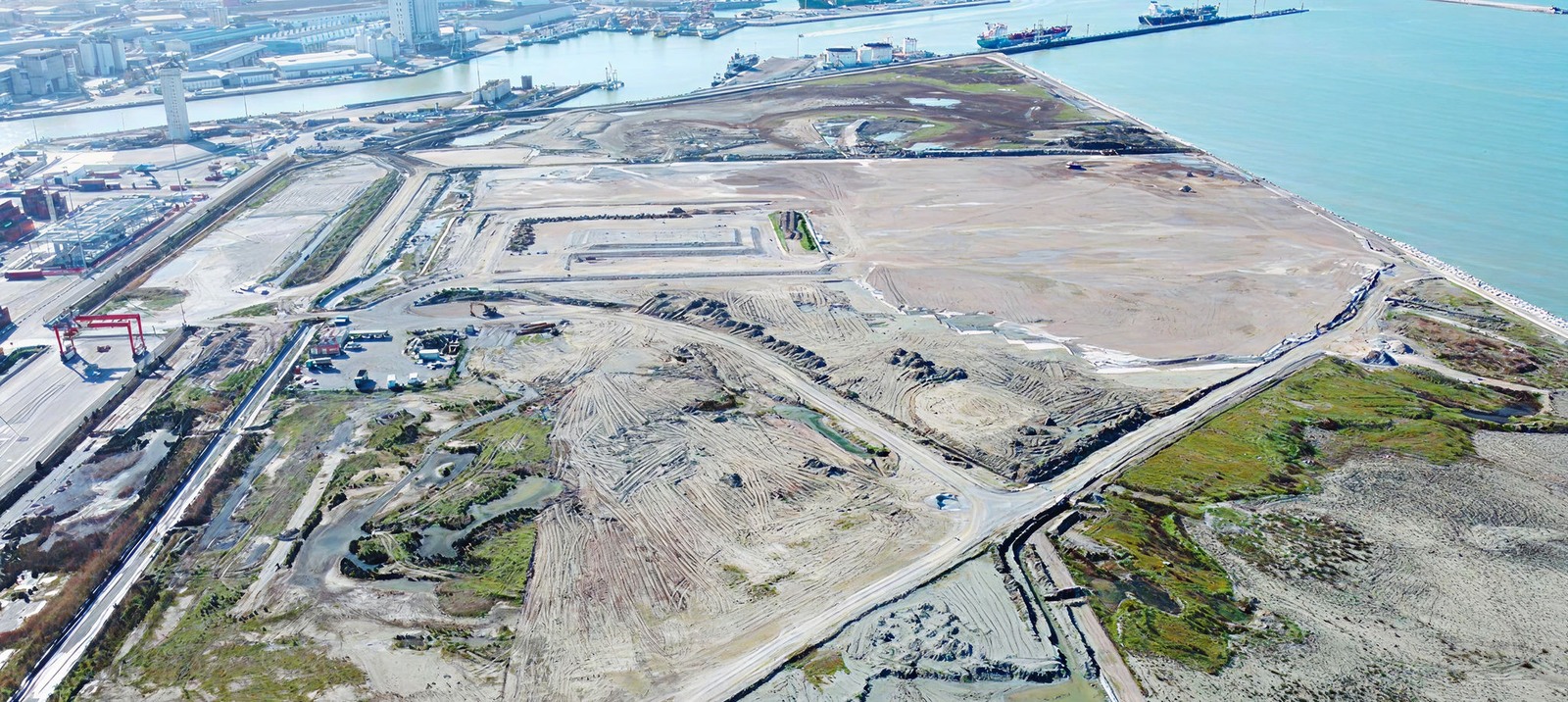 An expansive aerial view of a construction site near a coastline. The image showcases cleared land, industrial equipment, and surrounding water bodies. The setting includes docks and ships visible in the background, indicating proximity to a port. The scene highlights development activity in a coastal area.