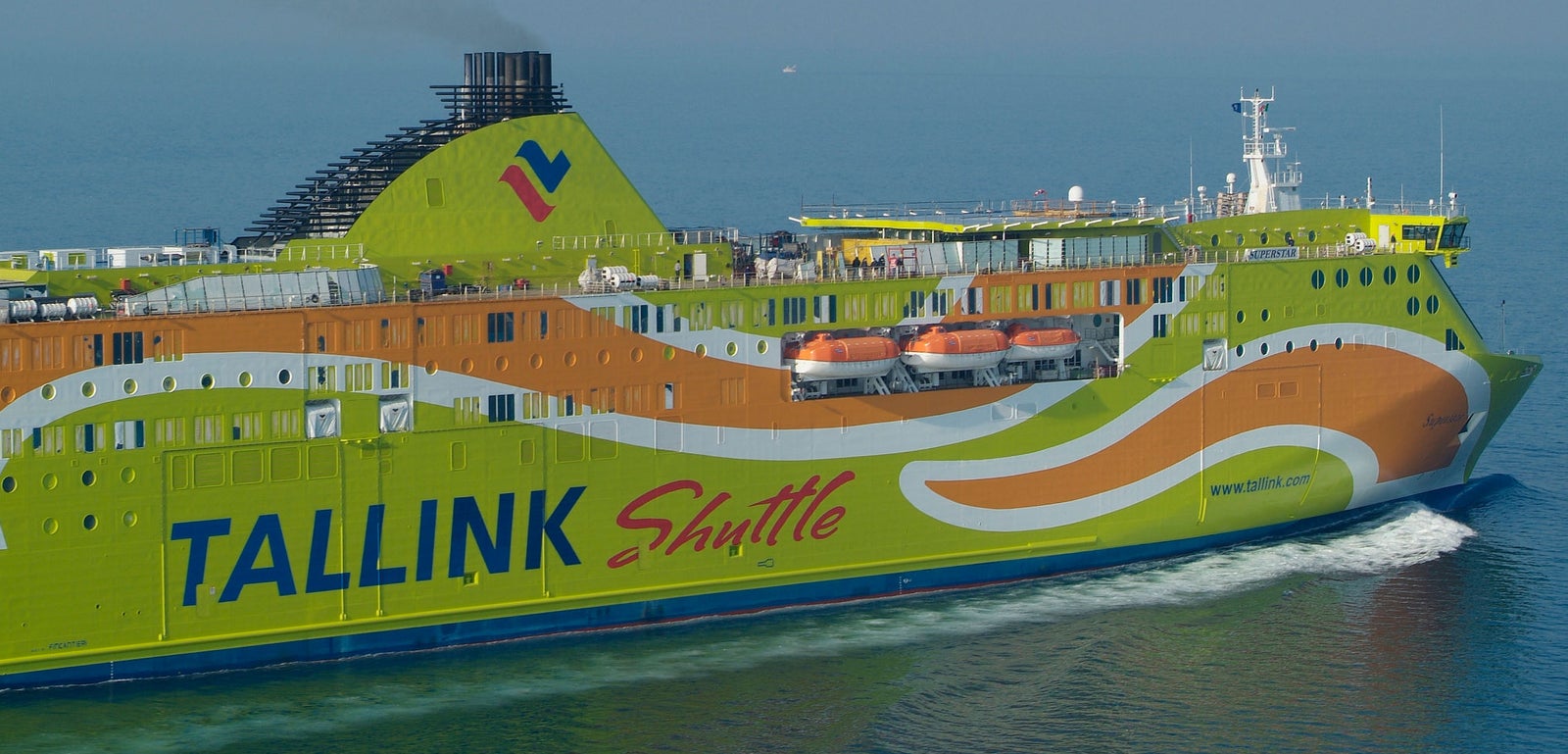 Fincantieri | Group - Business - Products - Ferries - Superstar: Vibrant Tallink Shuttle ferry in operation, colorful design, visible "Tallink Shuttle" text, lifeboats on deck, light smoke from chimney, cruising through calm waters, clear sky.