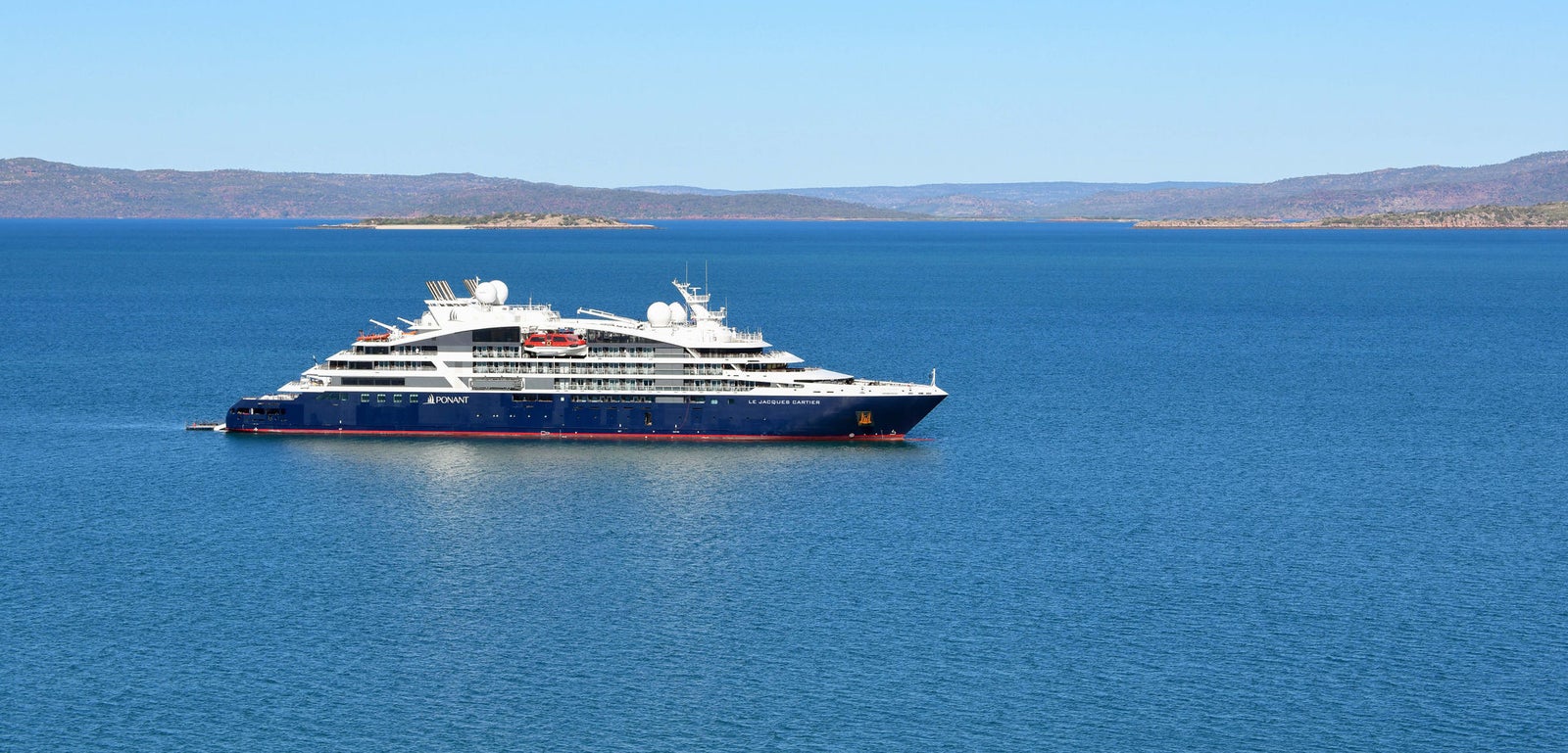 Fincantieri | Gruppo - Business - Prodotti - Navi da Crociera - Le Dumont d'Urville: Vista laterale della nave Le Dumont d'Urville operativa in navigazione su mare aperto, scafo dal profilo lineare, ponti passeggeri multipli, scritte "Le Dumont d'Urville" visibili sullo scafo, cielo sereno.