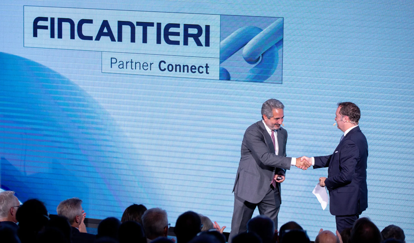 Fornitori e Partner – Fincantieri | Partnership: Due uomini in abiti formali si danno la mano sul palco durante l’evento Fincantieri Partner Connect, sfondo con logo Fincantieri, testo “Partner Connect” e grafica, pubblico visibile in primo piano, atmosfera professionale e corporate. 1° FIncantieri's Supplier Summit