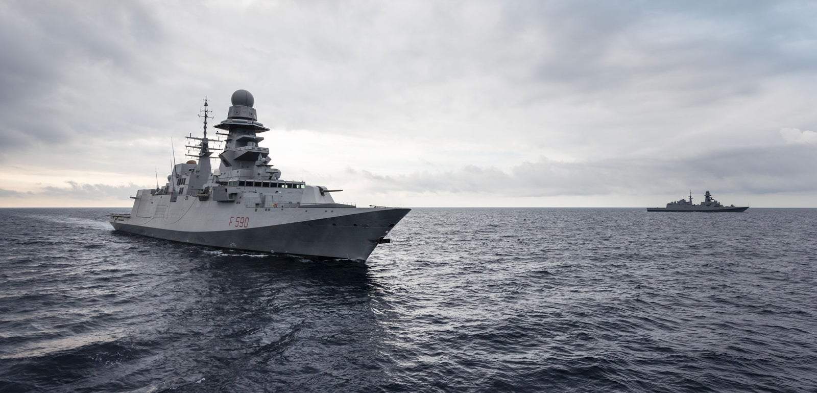 Fincantieri | Gruppo - Business - Prodotti - Navi per la difesa - Fregate - Classe Bergamini: Vista laterale di due navi operative in navigazione, primo piano con design moderno, radar e antenne visibili, seconda nave sullo sfondo.