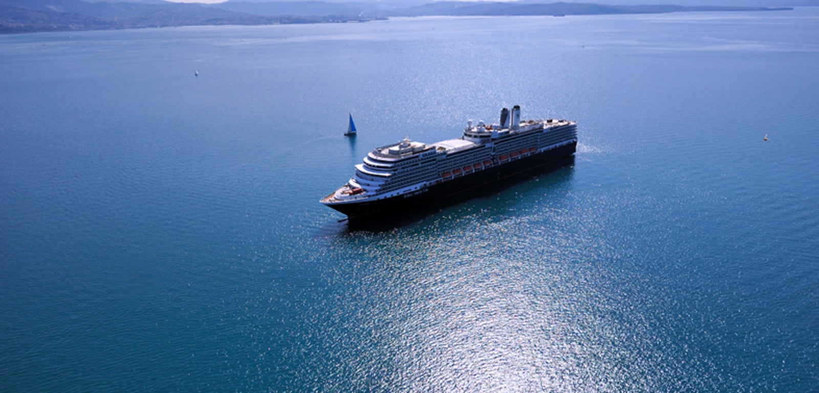  Fincantieri | Gruppo - Business - Prodotti - Navi da Crociera - Eurodam: Grande nave da crociera di lusso operativa che naviga su acque calme e aperte sotto cielo azzurro limpido, riflessi sereni di luce solare sull’acqua, piccola barca a vela visibile in lontananza.