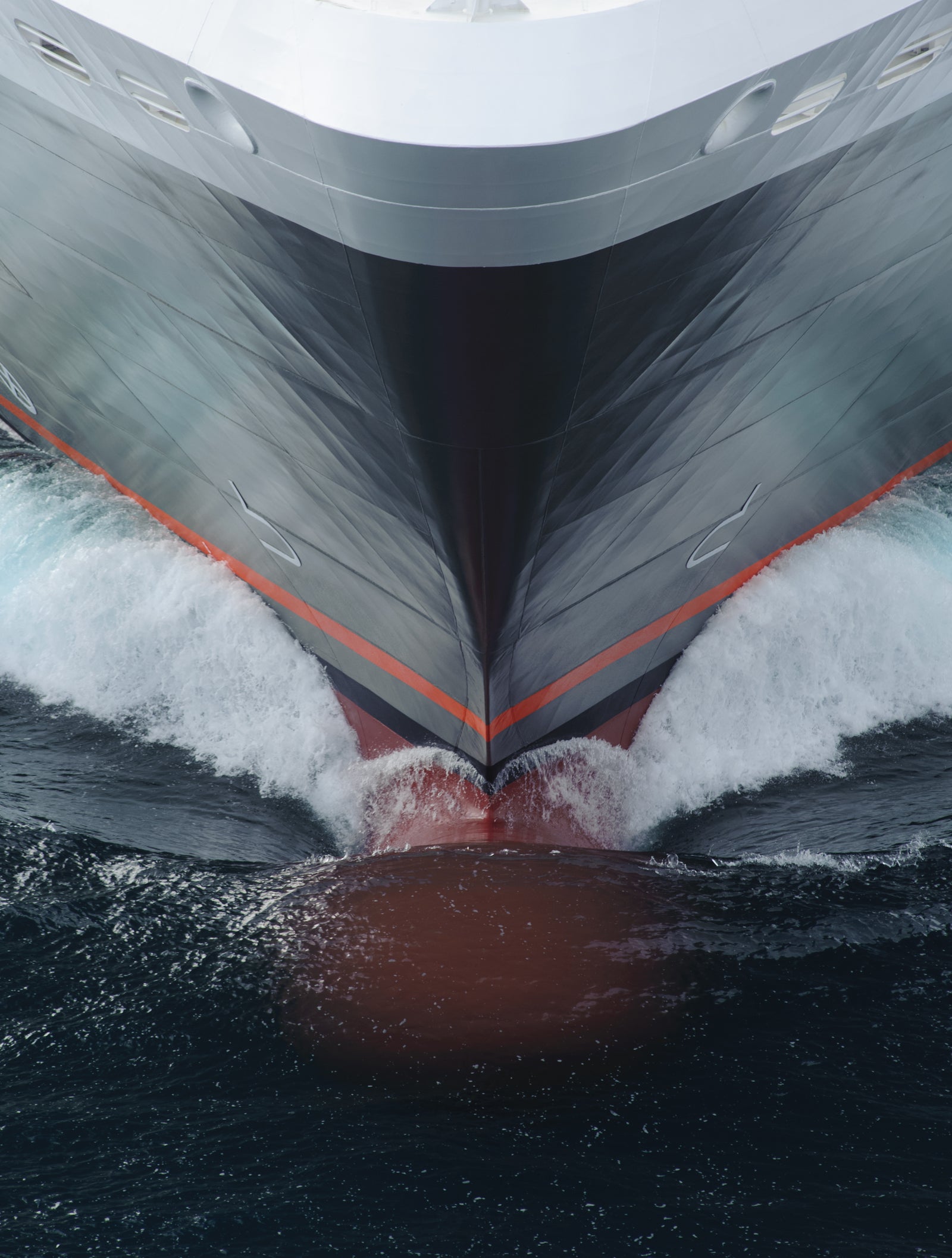 Fincantieri | Gruppo - Business - Prodotti - Navi da Crociera - Le Boreal: Vista ravvicinata della prua della nave operativa che fende le onde oceaniche, design slanciato con palette rosso e grigio, strisce arancioni a contrasto, movimento dinamico dell’acqua evidenzia potenza e velocità in ambiente marino.