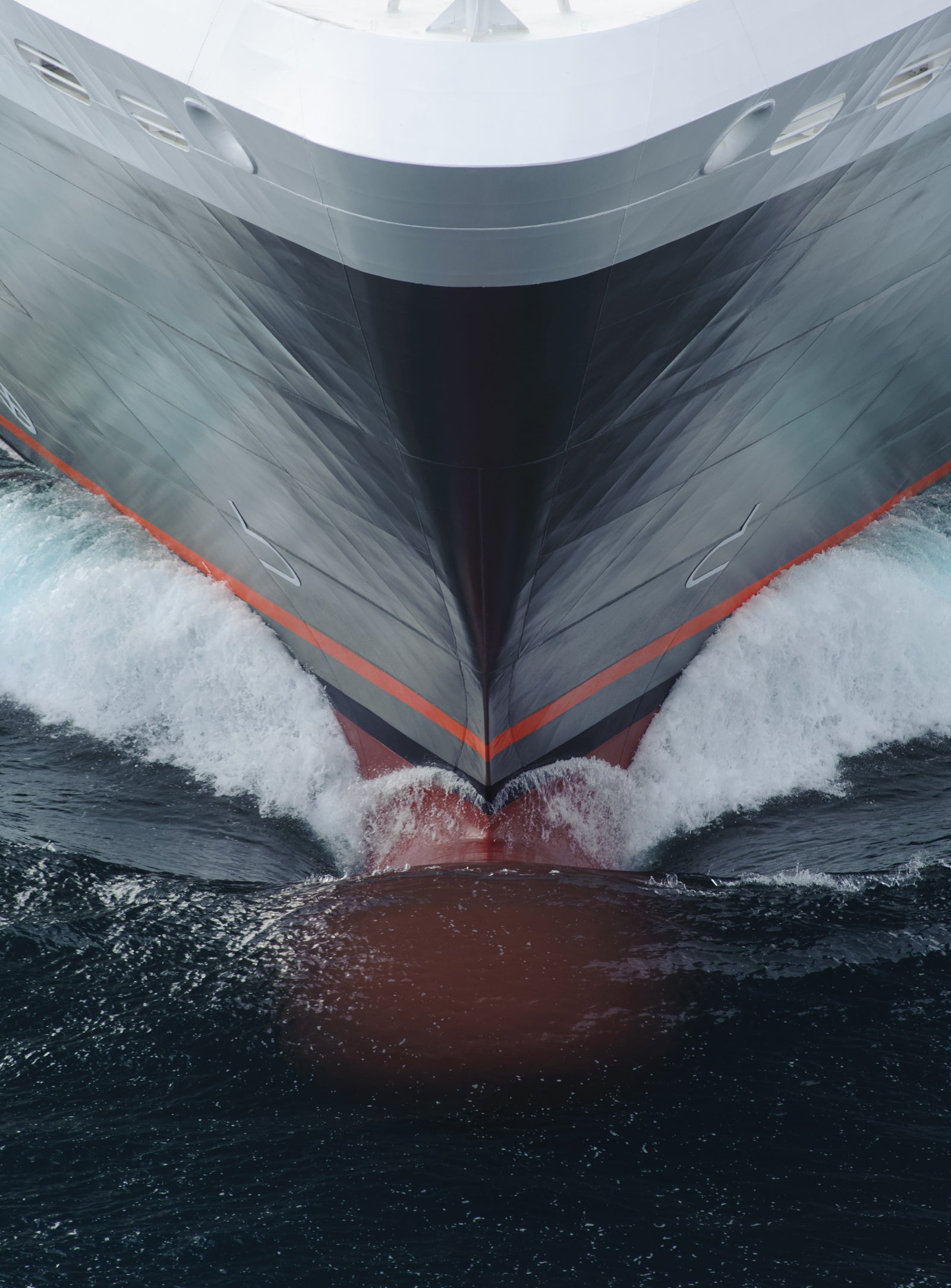  Fincantieri | Group - Business - Products - Cruise Ships - Le Boreal: Close-up view of ship’s bow in operation slicing through ocean waves, sleek design with red and gray color palette, orange stripes accent, dynamic water motion highlights power and speed in marine setting.