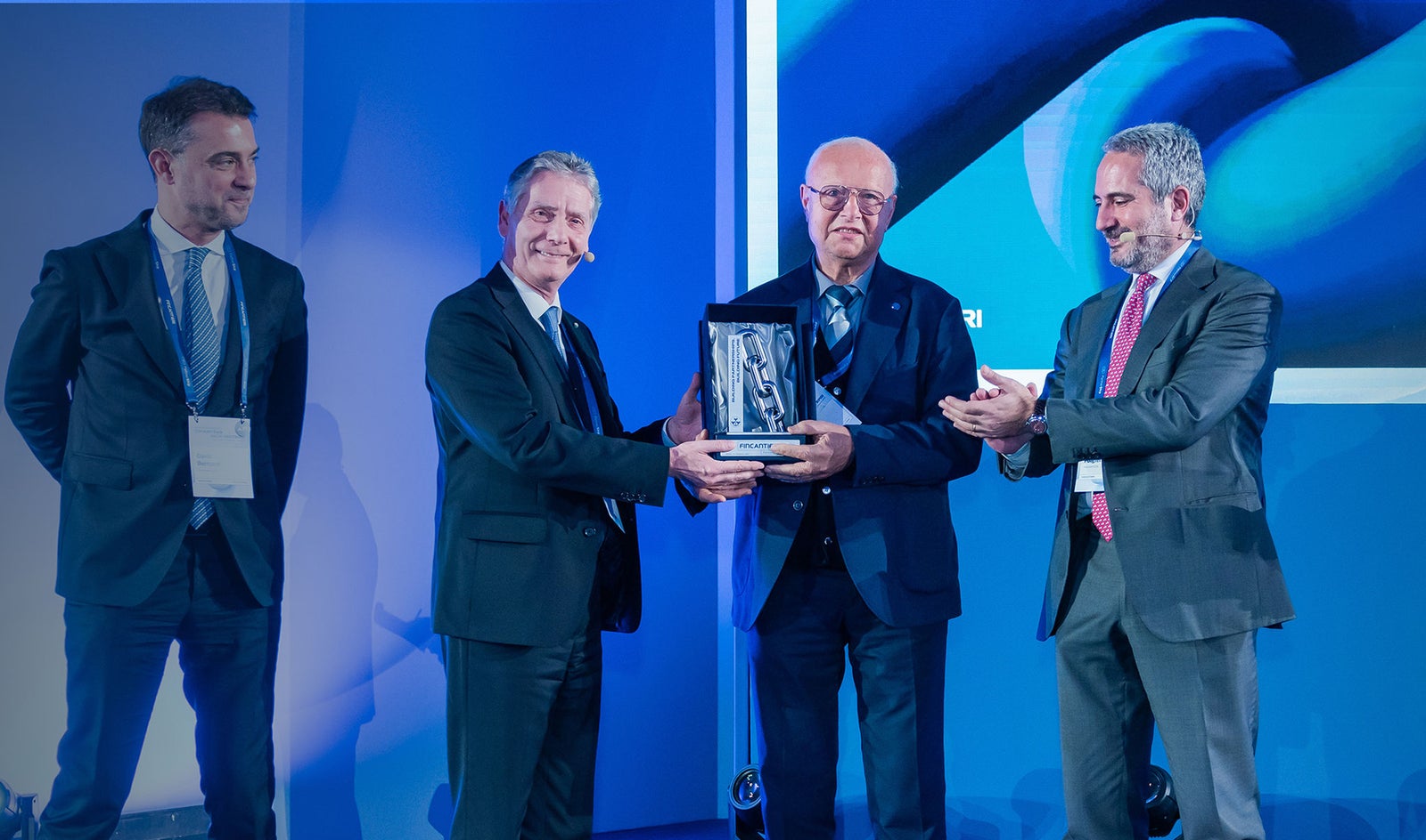 Fornitori e Partner – Fincantieri | Partnership: Gruppo di uomini in abiti formali su un palco durante una cerimonia di premiazione, uno tiene una targa o trofeo mentre gli altri applaudono, sfondo blu con visual astratte, ambiente professionale e corporate. 2° Fincantieri's Supplier Summit