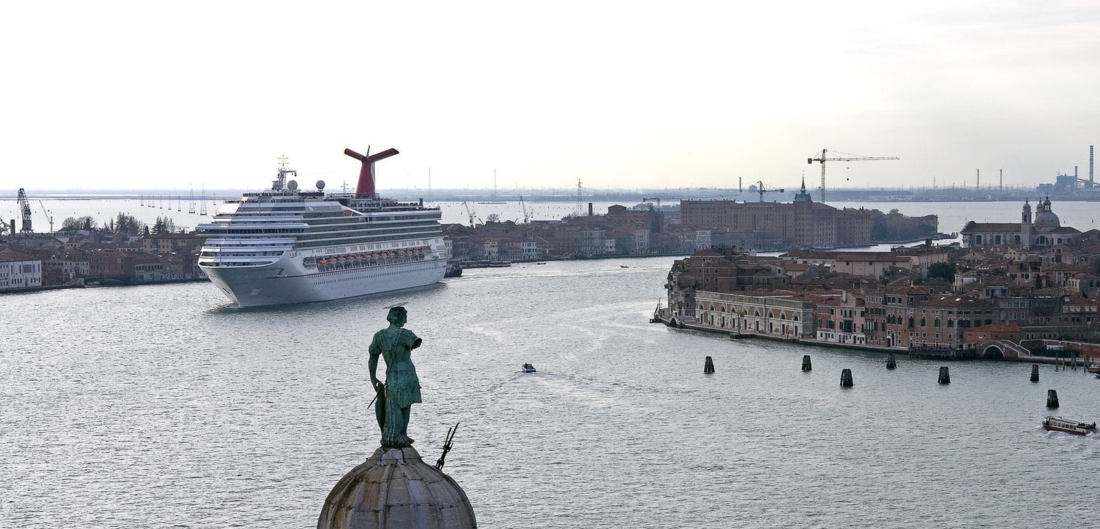  Fincantieri | Gruppo - Business - Prodotti - Navi da Crociera - Carnival Freedom: Grande nave da crociera operativa vista mentre si dirige verso Venezia, con architettura storica e canali in primo piano, skyline veneziano iconico con statue e tetti a cupola, fumaiolo rosso ben visibile in contrasto con i toni tenui del paesaggio urbano e dell’acqua.