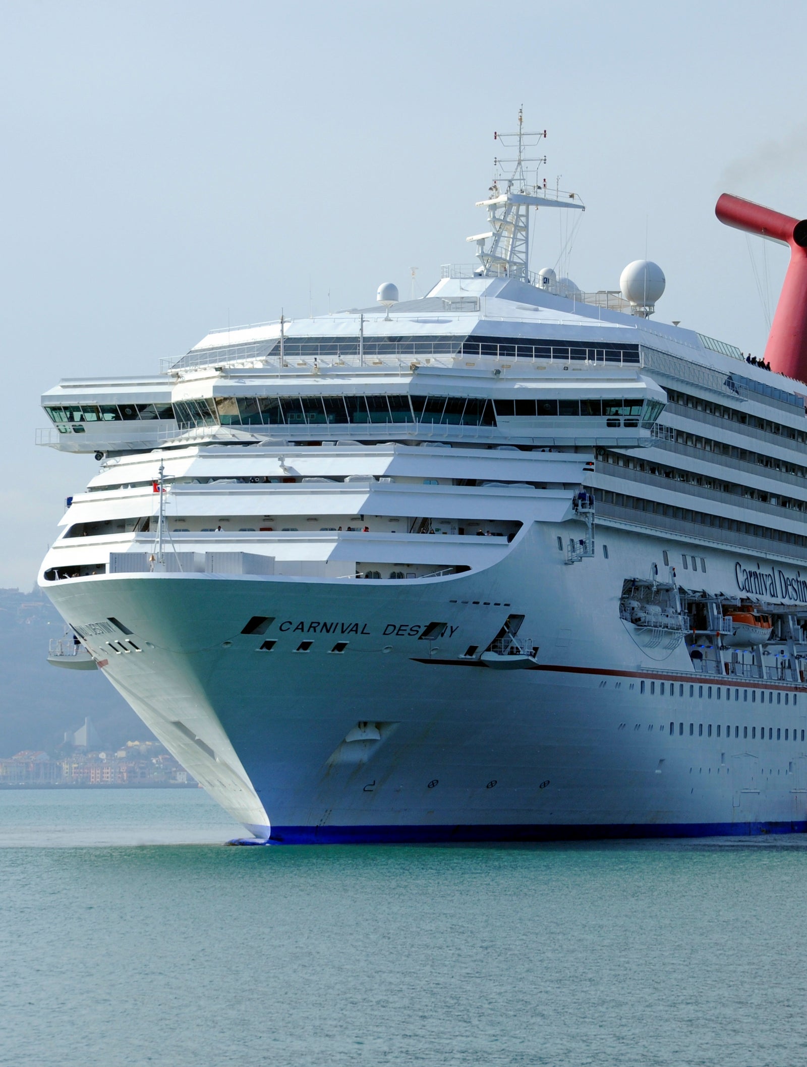 Fincantieri | Gruppo - Business - Prodotti - Navi da Crociera - Carnival Destiny: Nave da crociera bianca imponente etichettata 'Carnival Pride' in navigazione su acque calme, più ponti, scialuppe di salvataggio, fumaiolo rosso distintivo, linea di costa distante sotto cielo sereno.