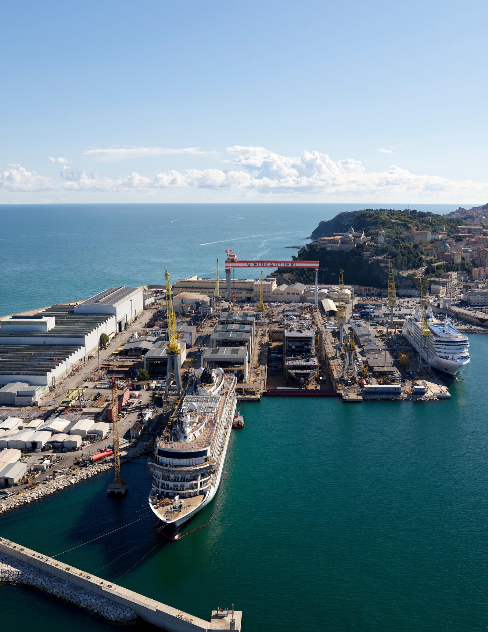 Fincantieri | Sostenibilità - Transizione Energetica: Large shipyard with several cruise ships under construction, visible cranes and industrial equipment.