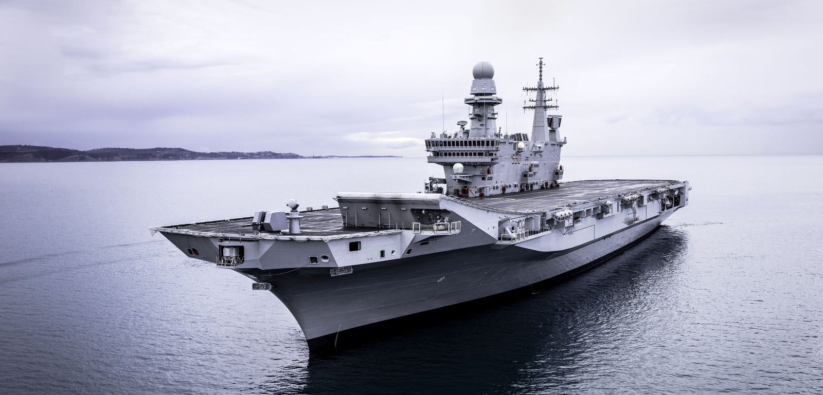 Fincantieri | Group - Business - Products - Naval Vessels - Aircraft Carriers and Amphibious Ships - Cavour: Large gray and white aircraft carrier "Cavour" in operation, long flight deck, prominent island structure, sailing on ocean, cloudy sky.