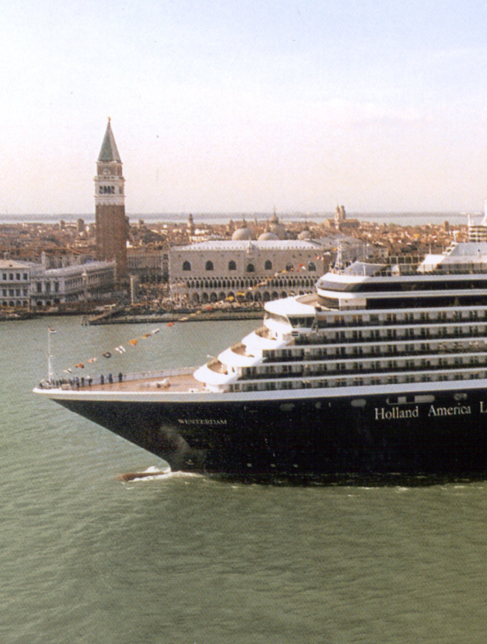 Fincantieri | Group - Business - Products - Cruise Ships - Noordam: Large cruise ship with dark hull in operation navigating the Venice lagoon, side profile view, bell tower and historic buildings lit by daylight in the background.