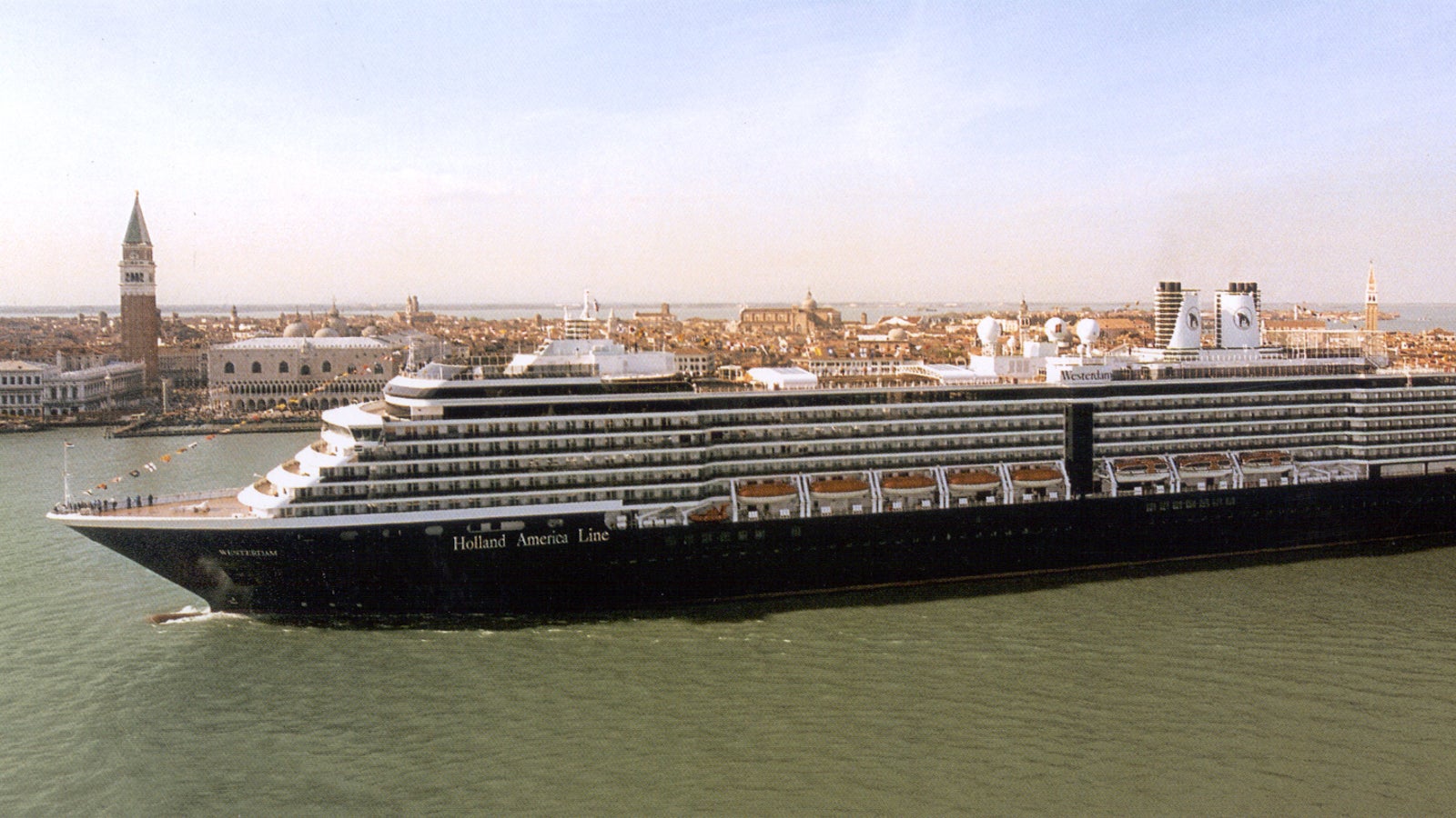 Fincantieri | Group - Business - Products - Cruise Ships - Noordam: Large cruise ship with dark hull in operation navigating the Venice lagoon, side profile view, bell tower and historic buildings lit by daylight in the background.