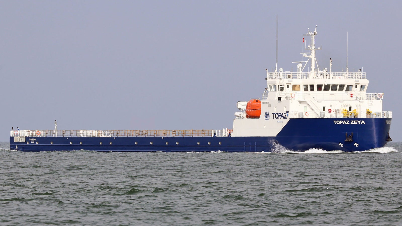 Prodotto - Nave Cargo Topaz Zeya: Vista laterale della nave operativa in navigazione, scafo blu e bianco con testo TOPAZ ZEYA visibile, ponte carico, scialuppa arancione montata lateralmente.