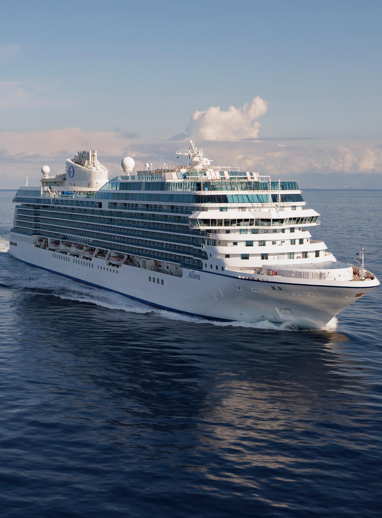 Fincantieri | Group - Companies - Subsidiaries and Affiliates - Fincantieri Services USA: Grey naval ship operationally sailing on calm waters at sunset, equipped with antennas, cranes and prominent central deck; background with mountainous coast and partly cloudy sky, serene marine environment.