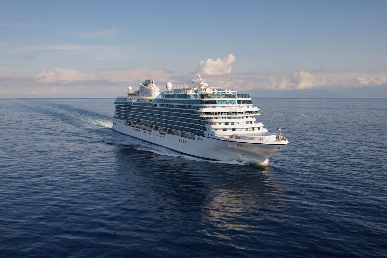 Fincantieri | Group - Companies - Subsidiaries and Affiliates - Fincantieri Services USA: Grey naval ship operationally sailing on calm waters at sunset, equipped with antennas, cranes and prominent central deck; background with mountainous coast and partly cloudy sky, serene marine environment.