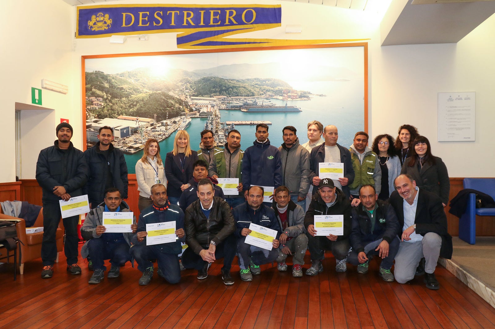 Newsroom - Comunicati stampa - 2026 - Fondazione Fincantieri e Società Dante Alighieri: al cantiere integrato di Riva Trigoso e Muggiano la consegna degli attestati dei corsi di lingua italiana per lavoratori stranieri: Gruppo di uomini e donne posano con attestati, ambiente interno con grande murale del cantiere navale, contesto formale di consegna.