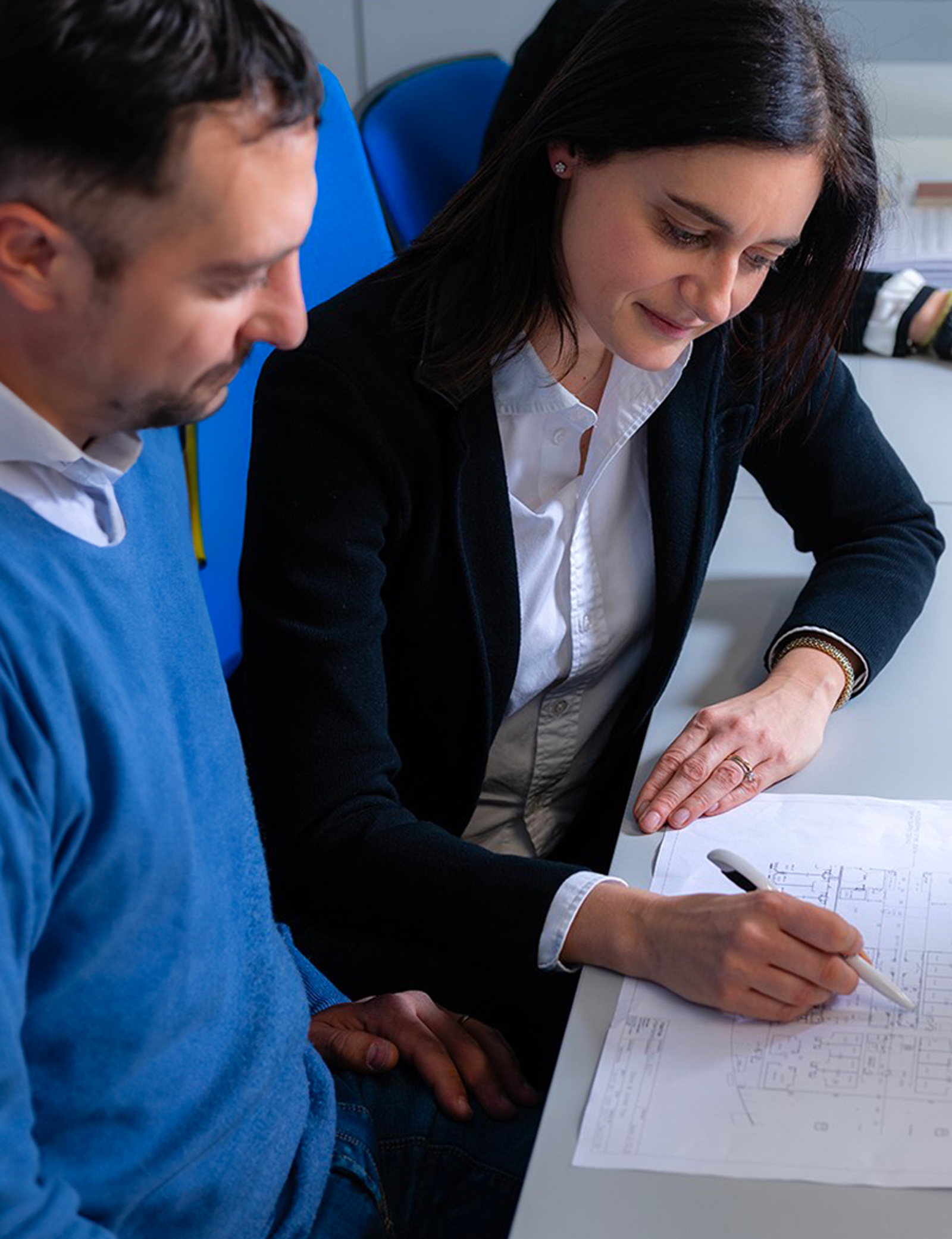 Fincantieri | Persone - Inclusione Vivere Fincantieri: Professionista in abbigliamento formale discute planimetrie architettoniche con collega in ufficio moderno, attenzione a precisione, collaborazione e dettagli progettuali, arredi blu e scrivania bianca.