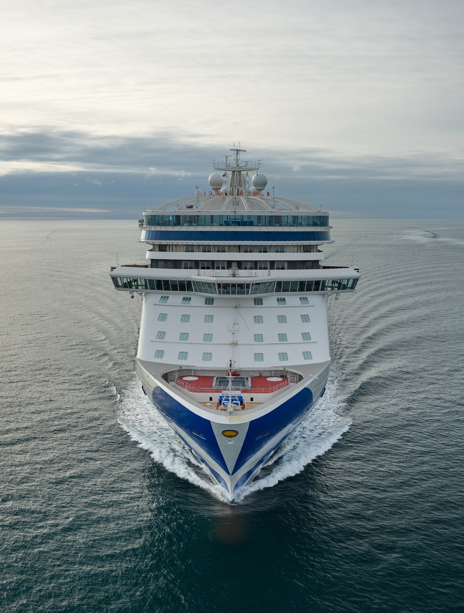Fincantieri | Group - Business - Products - Cruise Ships - Discovery Princess: Aerial front view of Discovery Princess cruise ship in operation, sailing on calm open waters under cloudy sky, blue and white design, visible decks and windows.