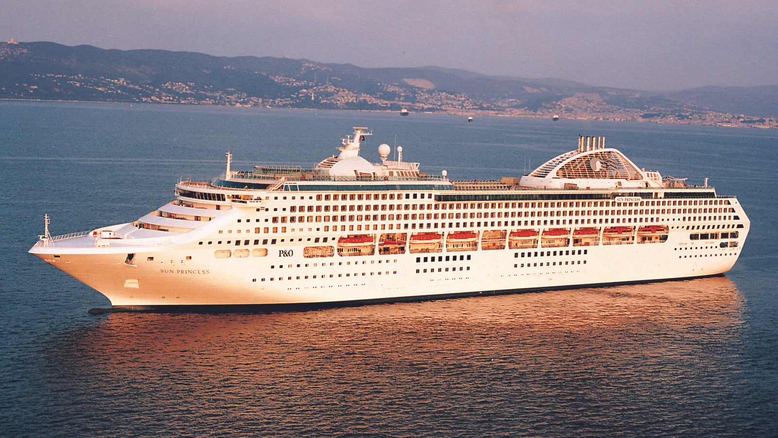 Fincantieri | Group - Business - Products - Cruise Ships - Sun Princess: Side view of Sun Princess cruise ship in operation navigating at sunset, spacious white hull, multiple decks, large windows, distinctive dome, coastal background.