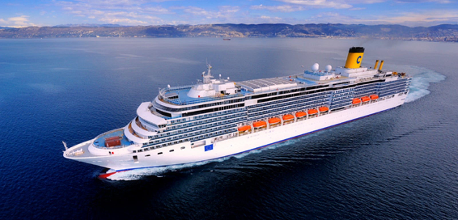 Fincantieri | Gruppo - Business - Prodotti - Navi da Crociera - Costa Luminosa: Grande nave da crociera di lusso operativa che scivola su acque calme e aperte sotto cielo blu limpido, più ponti, scialuppe, camino giallo distintivo con logo 'C' visibile, costa appena visibile sullo sfondo, scenario marittimo sereno.