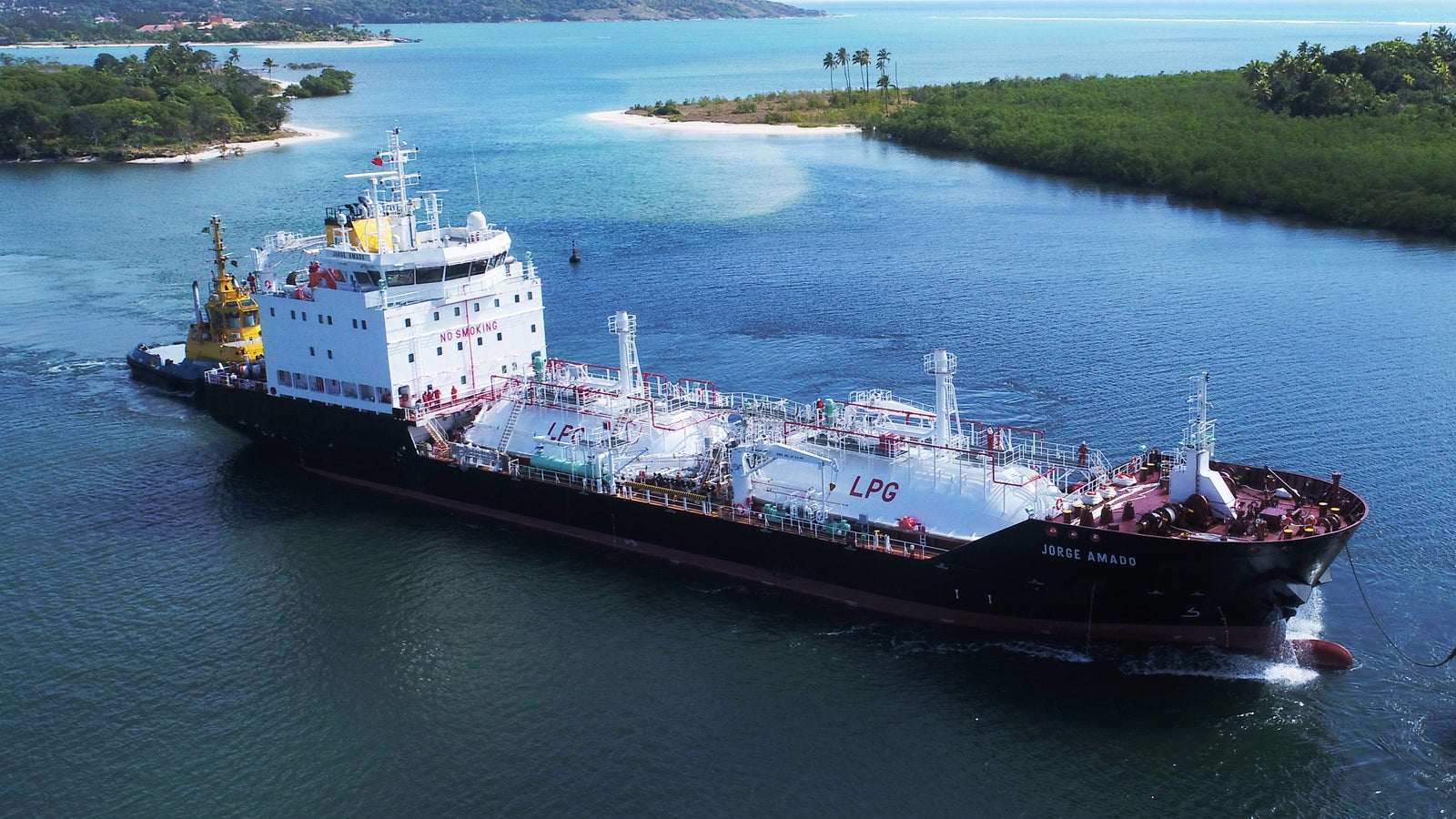 Prodotto - LPG Tanker Jorge Amado: Vista laterale della nave operativa in navigazione, testo LPG visibile sullo scafo, assistenza di rimorchiatore giallo, sfondo con isole tropicali verdi e mare calmo.