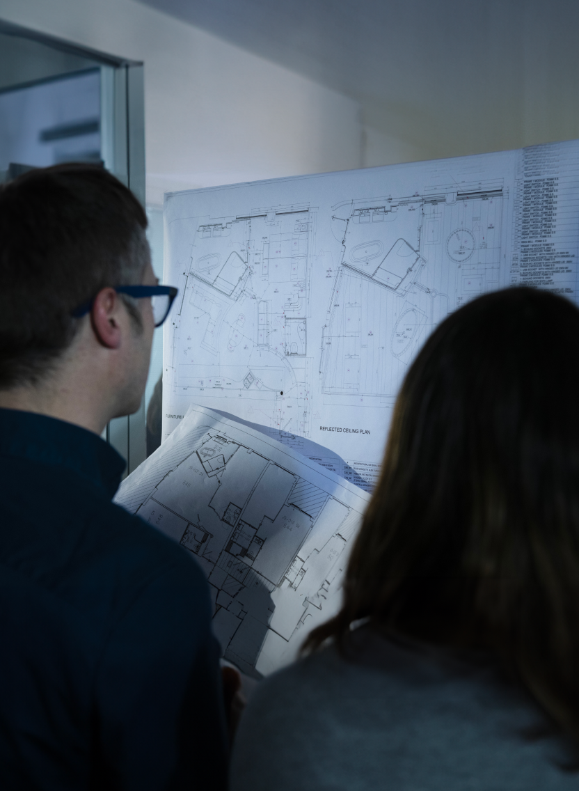 Fincantieri | People - Training and Talent: Two people, a man with glasses and a woman, review detailed architectural blueprints in a bright indoor setting, collaborating on technical drawings and design activities.