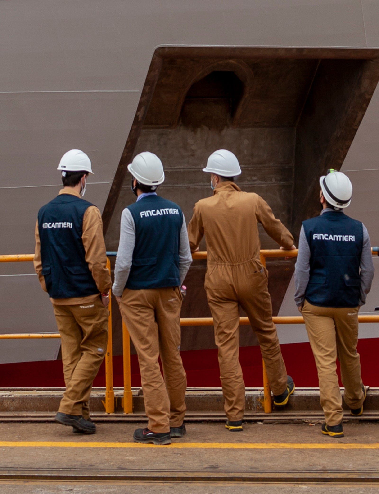 Fincantieri | About Us - Future on Board: Group of workers wearing safety helmets and uniforms near the hull of a large white ship with a red lower stripe, in a port or shipyard environment with safety railings, workers engaged in observation or inspection activities.