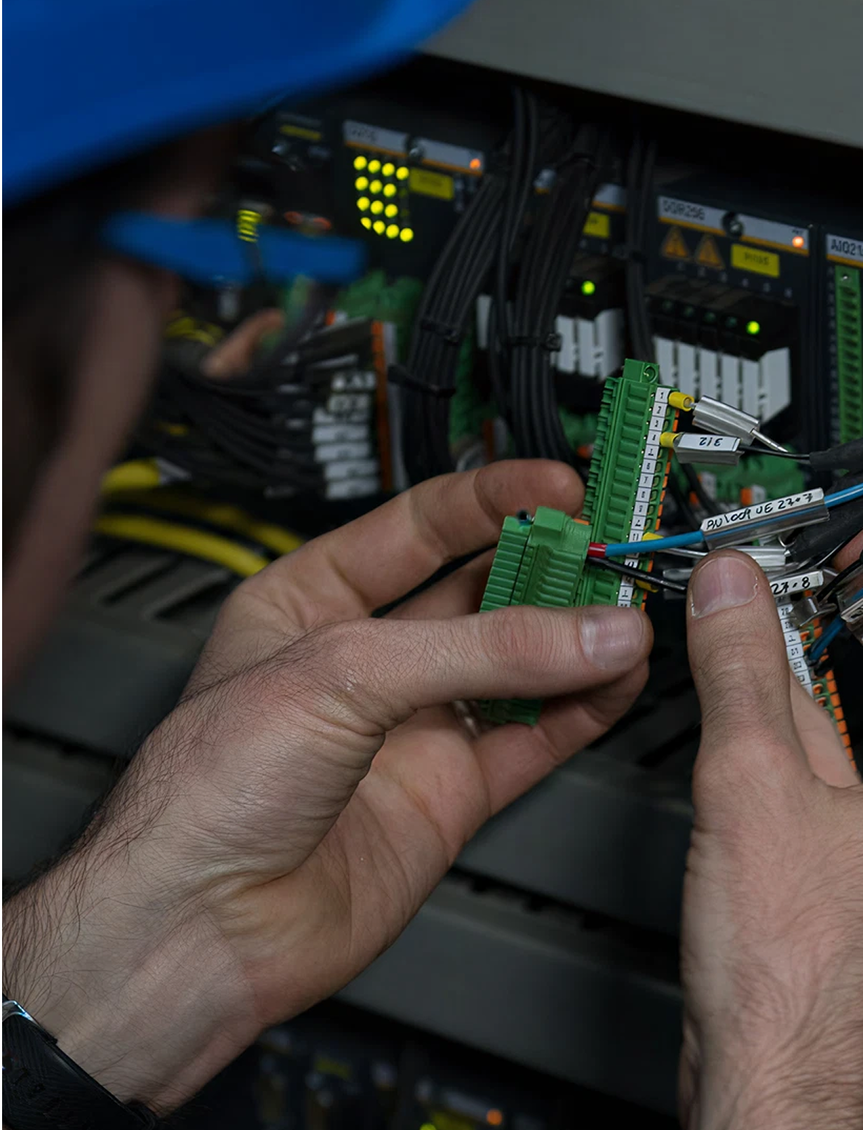 Fincantieri | Group - Subsidiaries and Affiliates - Fincantieri SI: Close-up of technician’s hands connecting wires to a green terminal block in an industrial electrical panel; control room setting, visible cables and illuminated indicators, technician in blue safety helmet for professional environment.
