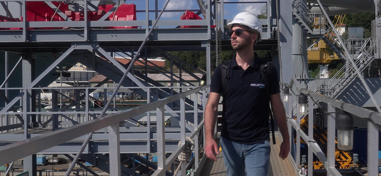 Fincantieri – People: Person wearing a white helmet and black polo shirt walking on a metal platform in an industrial environment, with steel structures, machinery, and blue sky in the background; operational context of a shipyard or production facility.