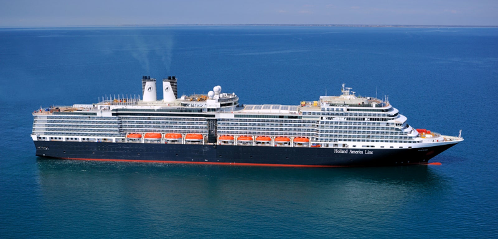 Fincantieri | Gruppo - Business - Prodotti - Navi da Crociera - Eurodam: Grande nave della Holland America Line operativa che naviga su acque calme e blu sotto cielo sereno, più ponti, scialuppe di salvataggio, due fumaioli gemelli con fumo chiaro, nome chiaramente visibile sul lato.