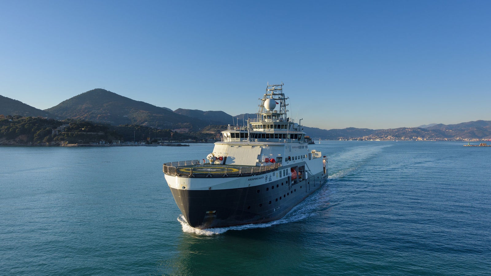 Prodotto - Nave da Ricerca Polare: Vista laterale della nave operativa in navigazione, struttura dettagliata, sfondo collinare e costiero, ambiente marittimo sereno.