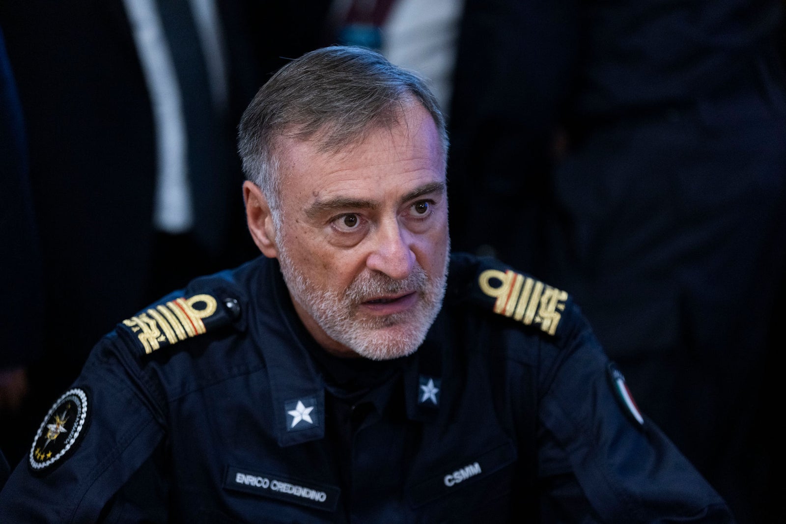 Fincantieri | Newsroom, Deep: A military officer is seen wearing a formal navy blue uniform adorned with insignias and badges. The setting appears to be indoors, possibly during an official event or gathering. Visible details include embroidered patches, stars, and text such as 'Enrico Credendino ' and 'CSMM' on the uniform.