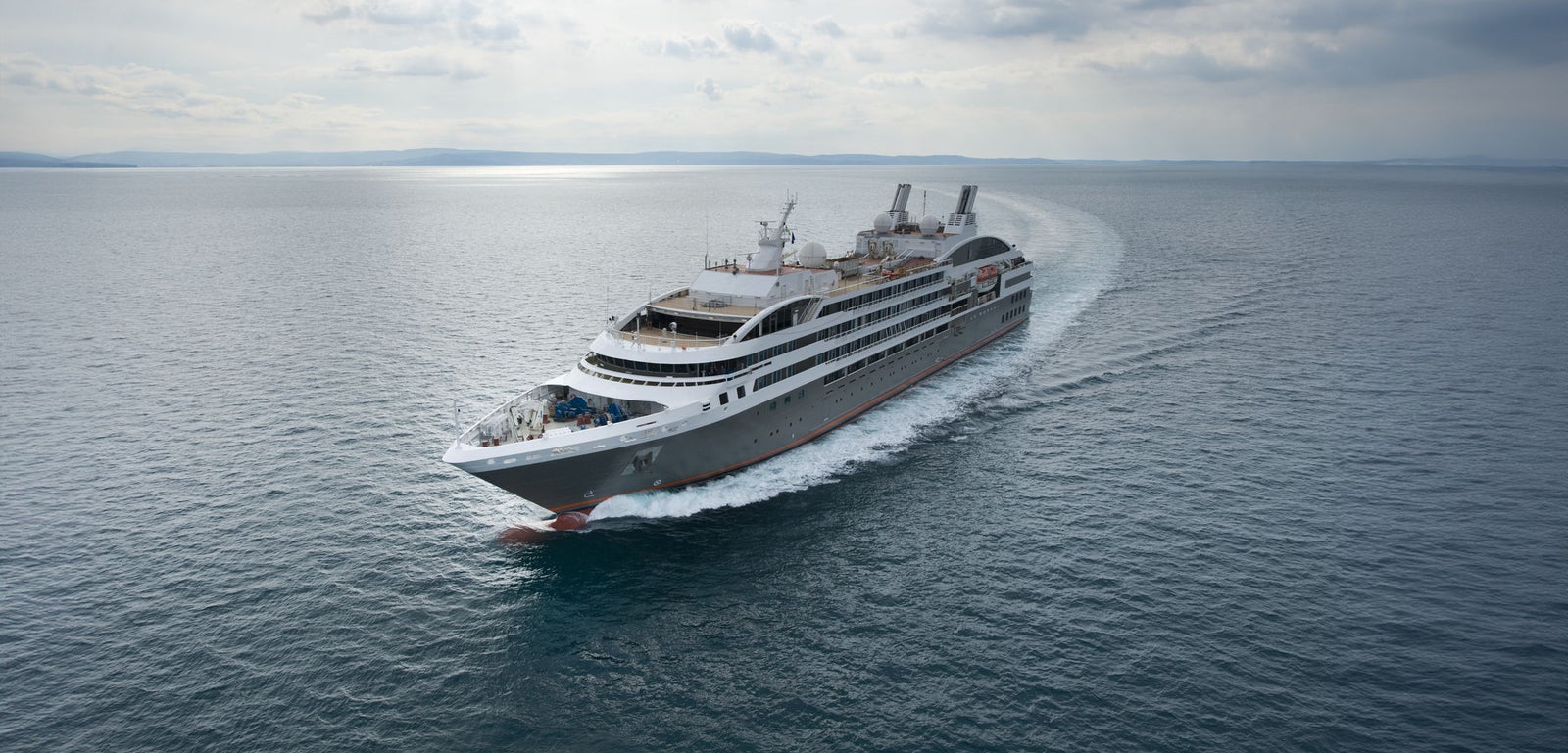 Fincantieri | Group - Business - Products - Cruise Ships - Le Boreal: View of Le Boreal cruise ship in operation sailing near coastline, visible white wake, multiple passenger decks, dark linear profile hull, distant hills in background.