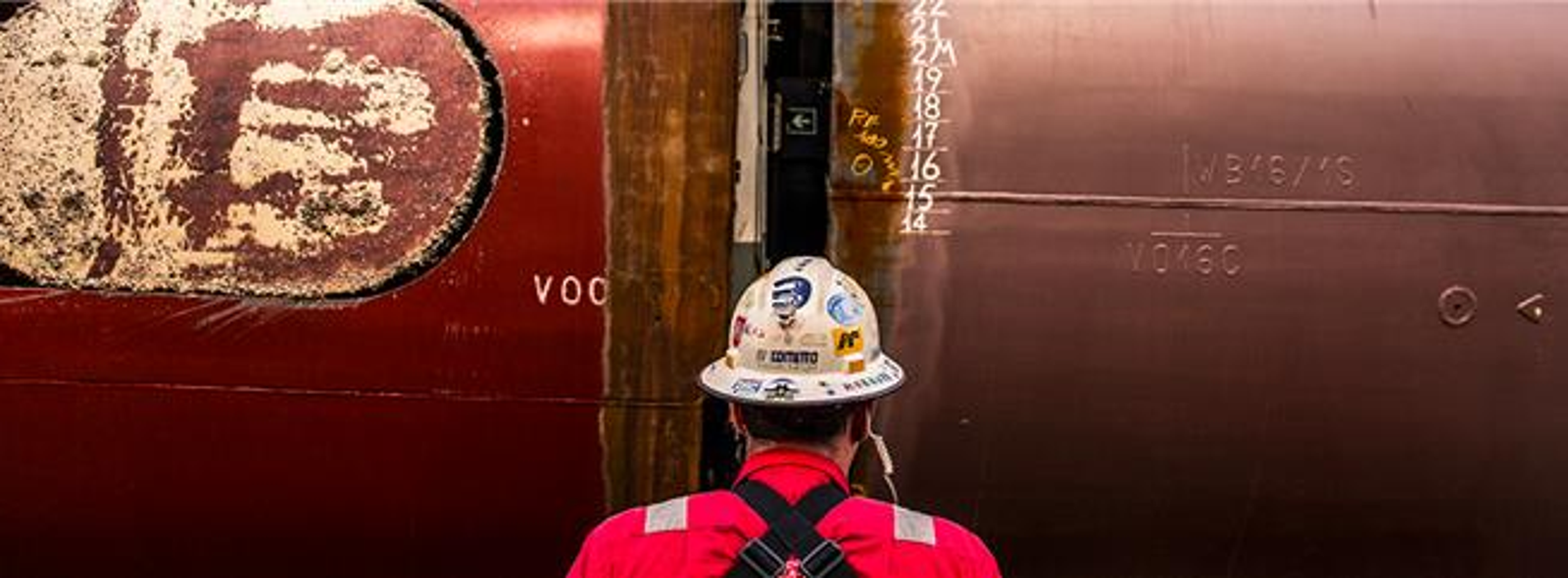 Servizio - Riparazioni e Trasformazioni Navali: Operatore con uniforme rossa e casco davanti a scafo nave in riparazione, segni di saldatura e numeri di misura (15 e 16) evidenti sulla superficie metallica, ambiente industriale.