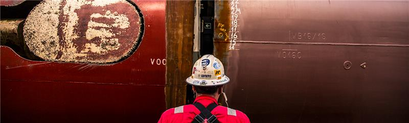 Fincantieri | Servizi - Riparazioni e Trasformazioni Navali: Operatore con uniforme rossa e casco davanti a scafo nave in riparazione, segni di saldatura e numeri di misura (15 e 16) evidenti sulla superficie metallica, ambiente industriale.