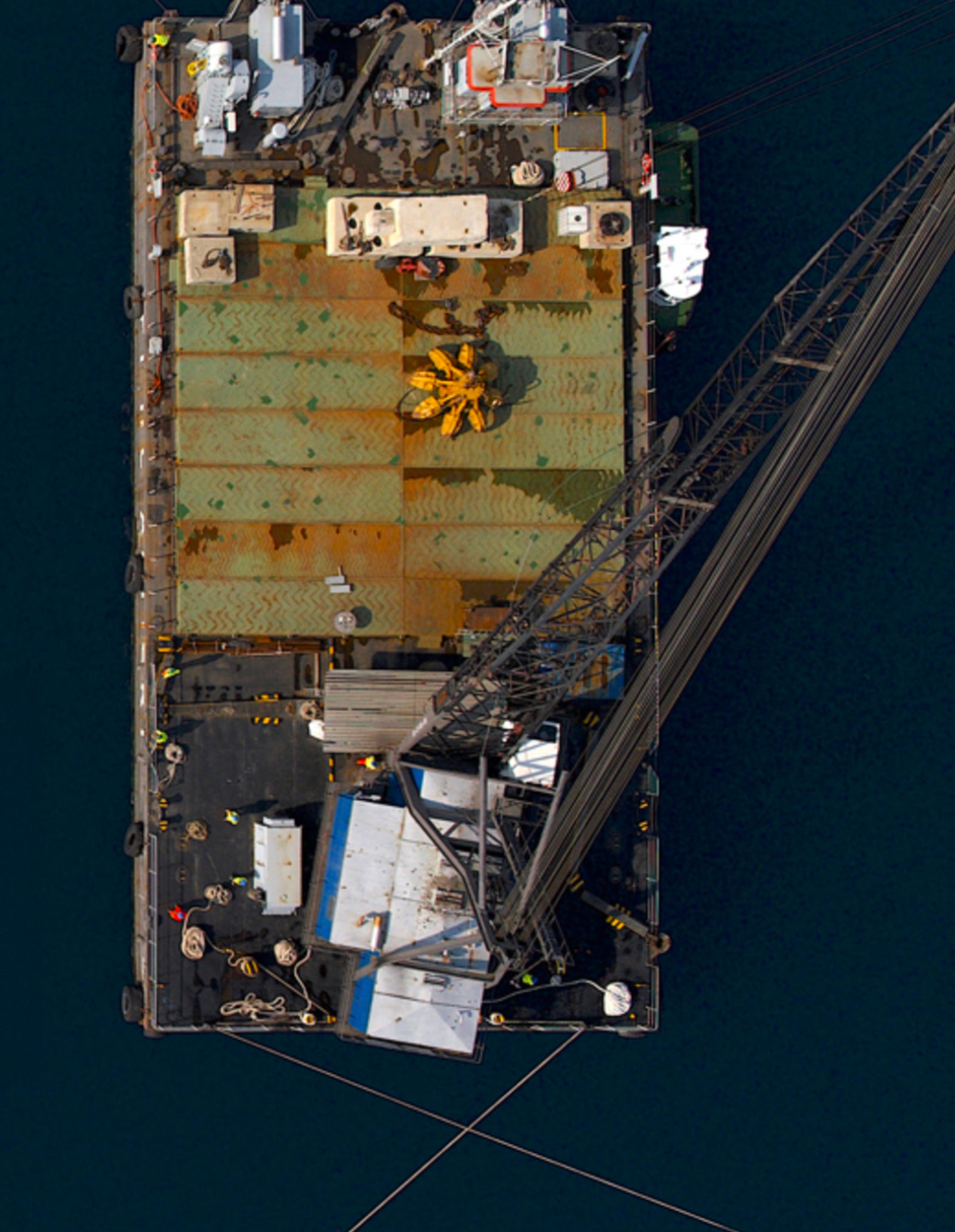 Fincantieri | Group - Subsidiaries and Affiliates - Fincantieri Opere Marittime: Top-down view of a large industrial barge equipped with a crane floating on dark water; deck features rusted metal panels, machinery, and yellow crane arm, suggesting maritime or construction activity.