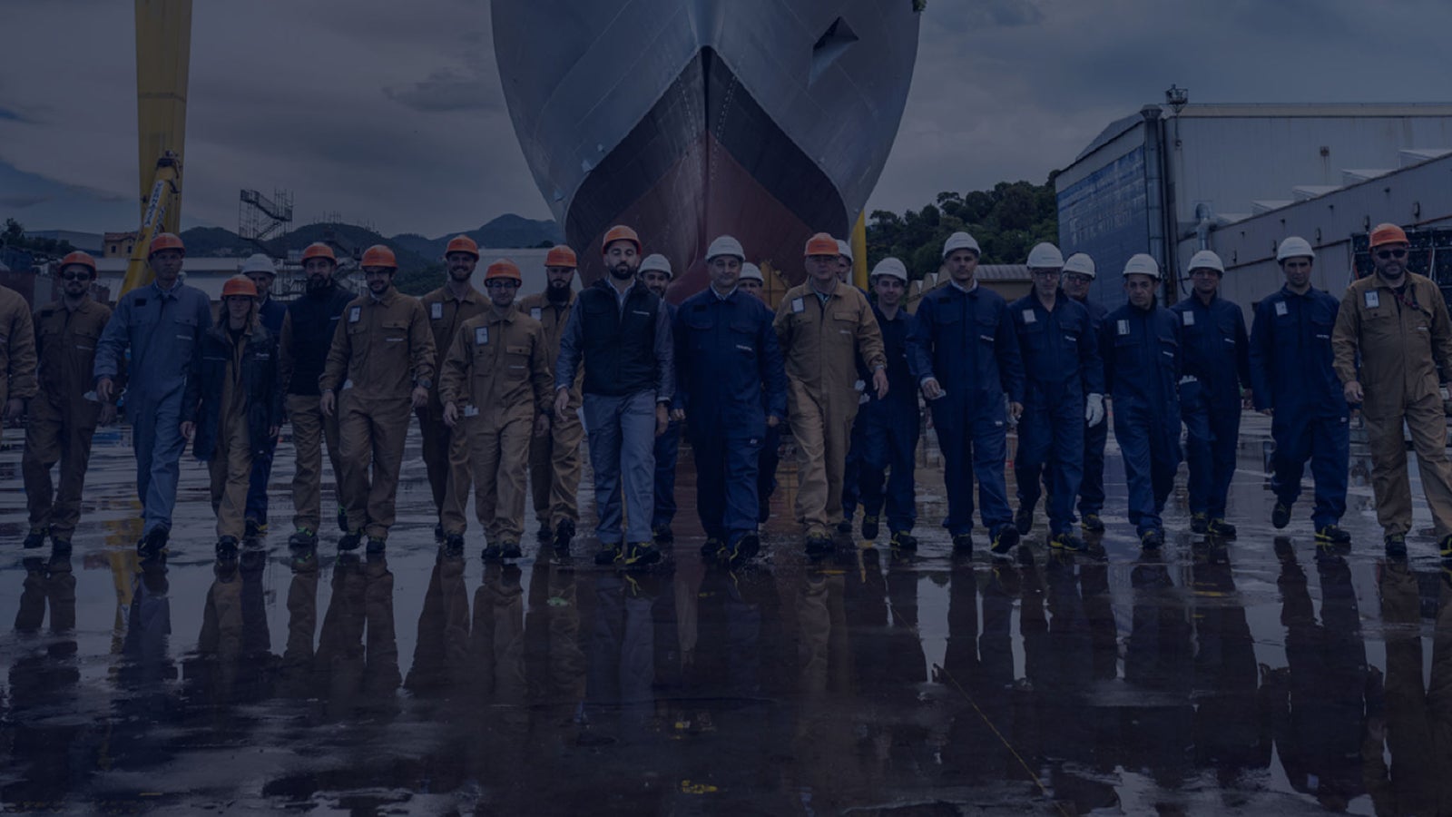 Fincantieri | Persone - Lavora con Noi: Uomo con casco bianco e giacca con scritta Fincantieri cammina in grande impianto industriale, strutture metalliche e luce soffusa, contesto di produzione o cantieristica. Candidati