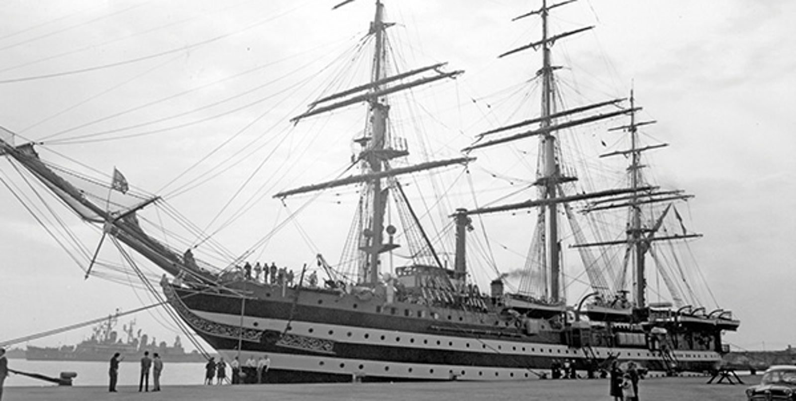 Fincantieri | About Us - Historic Ships: Large historic multi-masted sailing ship docked in port, black and white photo highlighting rigging and classic design, people near the ship for context and scale, another ship and calm waters in the background.