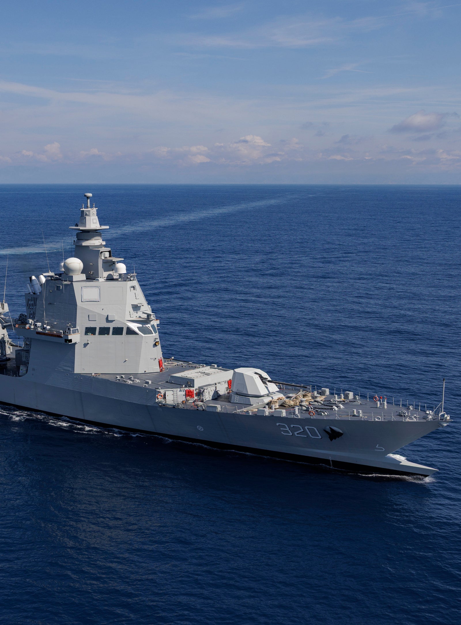 Fincantieri | Group - Companies - Subsidiaries and Affiliates - Orizzonte Sistemi Navali: Modern operational military ship sailing on deep blue ocean under a clear sky, equipped with advanced radars and armaments, distinctive wake emphasizing speed and agility; contrast between calm environment and the powerful presence of the ship.
