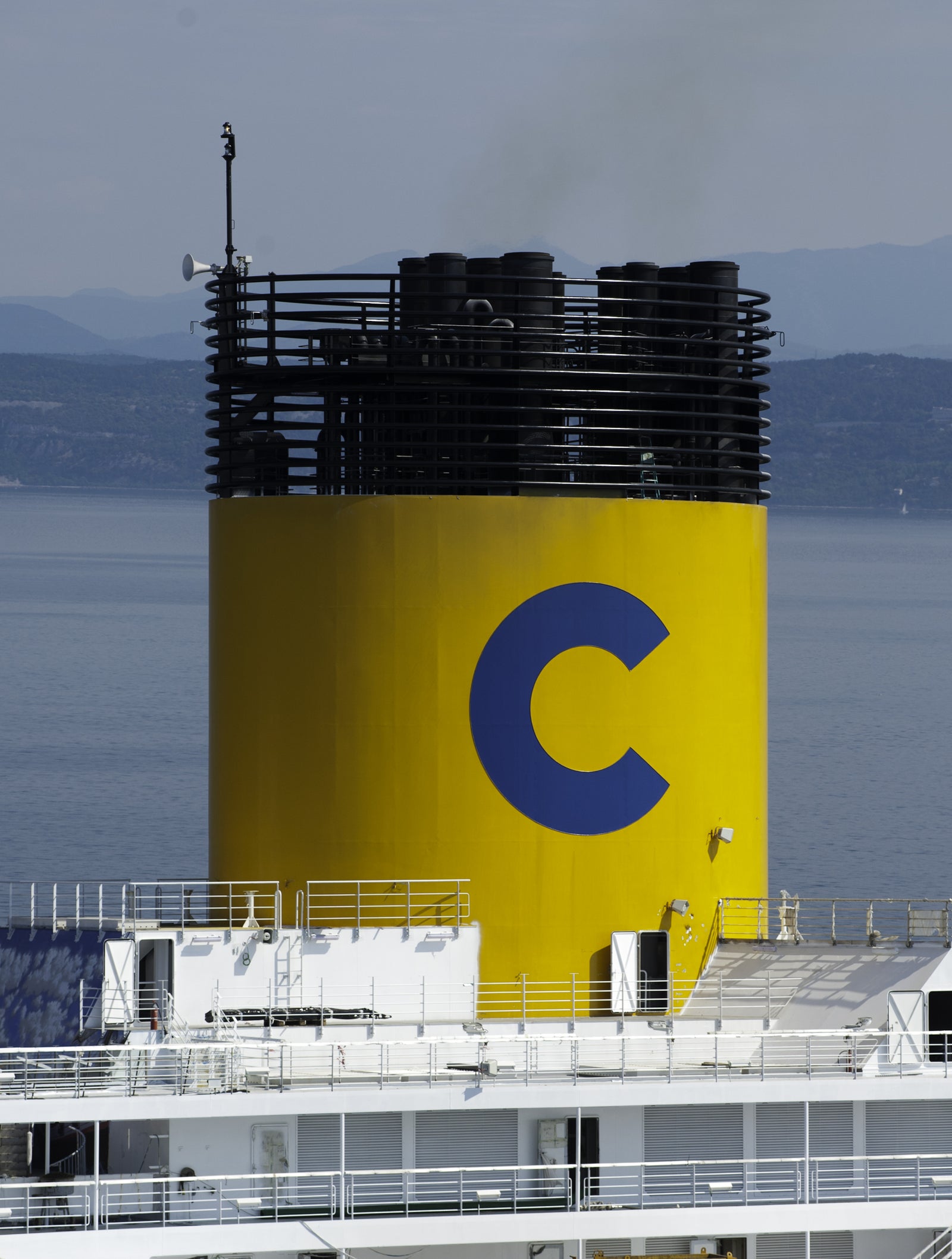 Fincantieri | Group - Business - Products - Cruise Ships - Costa Favolosa: Close-up view of ship’s bright yellow chimney with large blue 'C' logo, vessel docked near calm waterfront, distant mountains in background, industrial design contrasts with serene natural setting.
